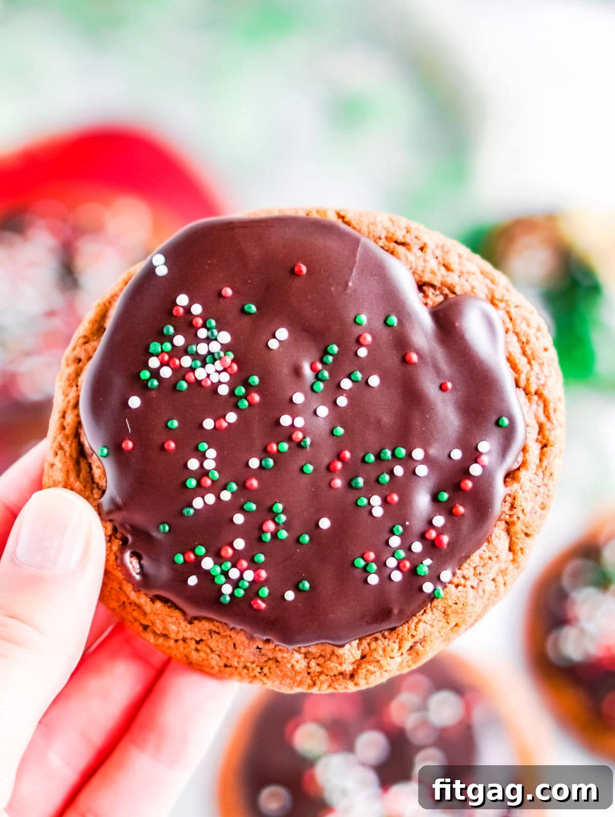 A hand holding a glazed chocolate cookie adorned with vibrant red, green, and white sprinkles, illuminated beautifully by natural light.