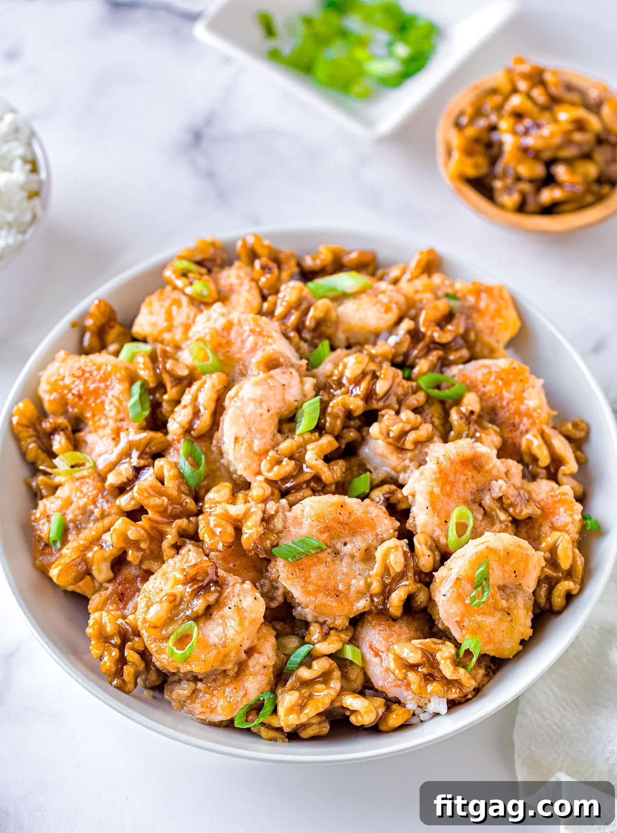 White bowl brimming with Honey Walnut Shrimp, with a small bowl of walnuts and a bunch of green onions in the soft-focus background.