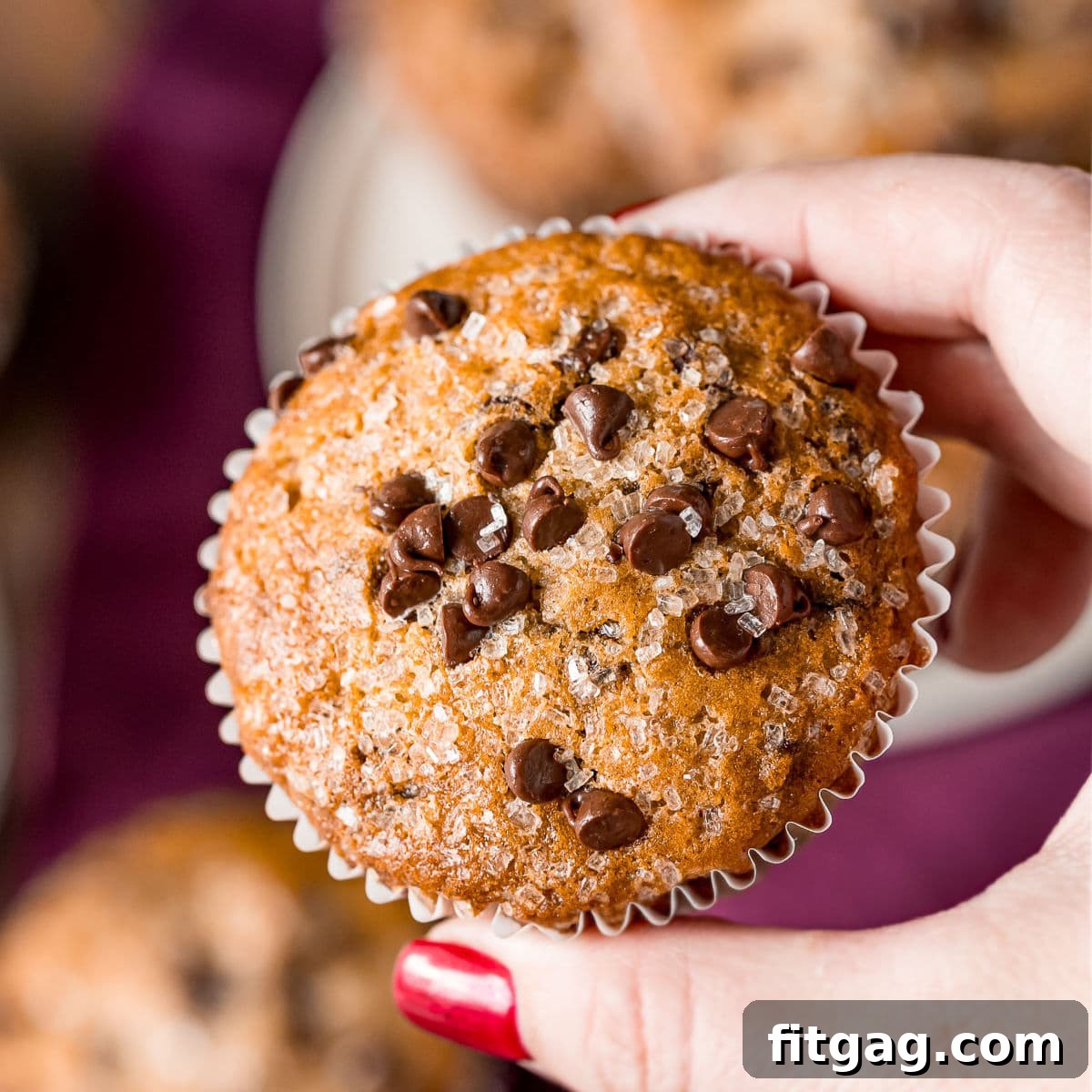 Effortless Chocolate Chip Muffins 6 A hand gently holding a freshly baked chocolate chip muffin aloft, with a plate of more delicious muffins blurred in the background.