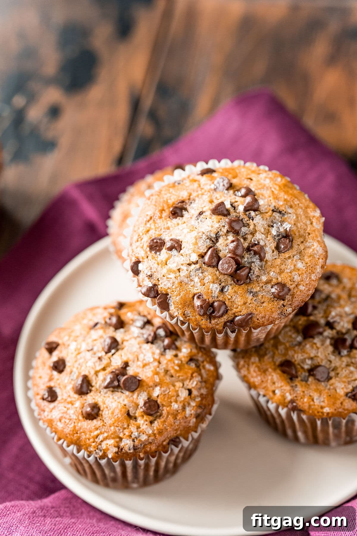 Effortless Chocolate Chip Muffins 9 Four perfectly baked chocolate chip muffins resting on a white plate, with a rich burgundy napkin positioned beneath the plate.