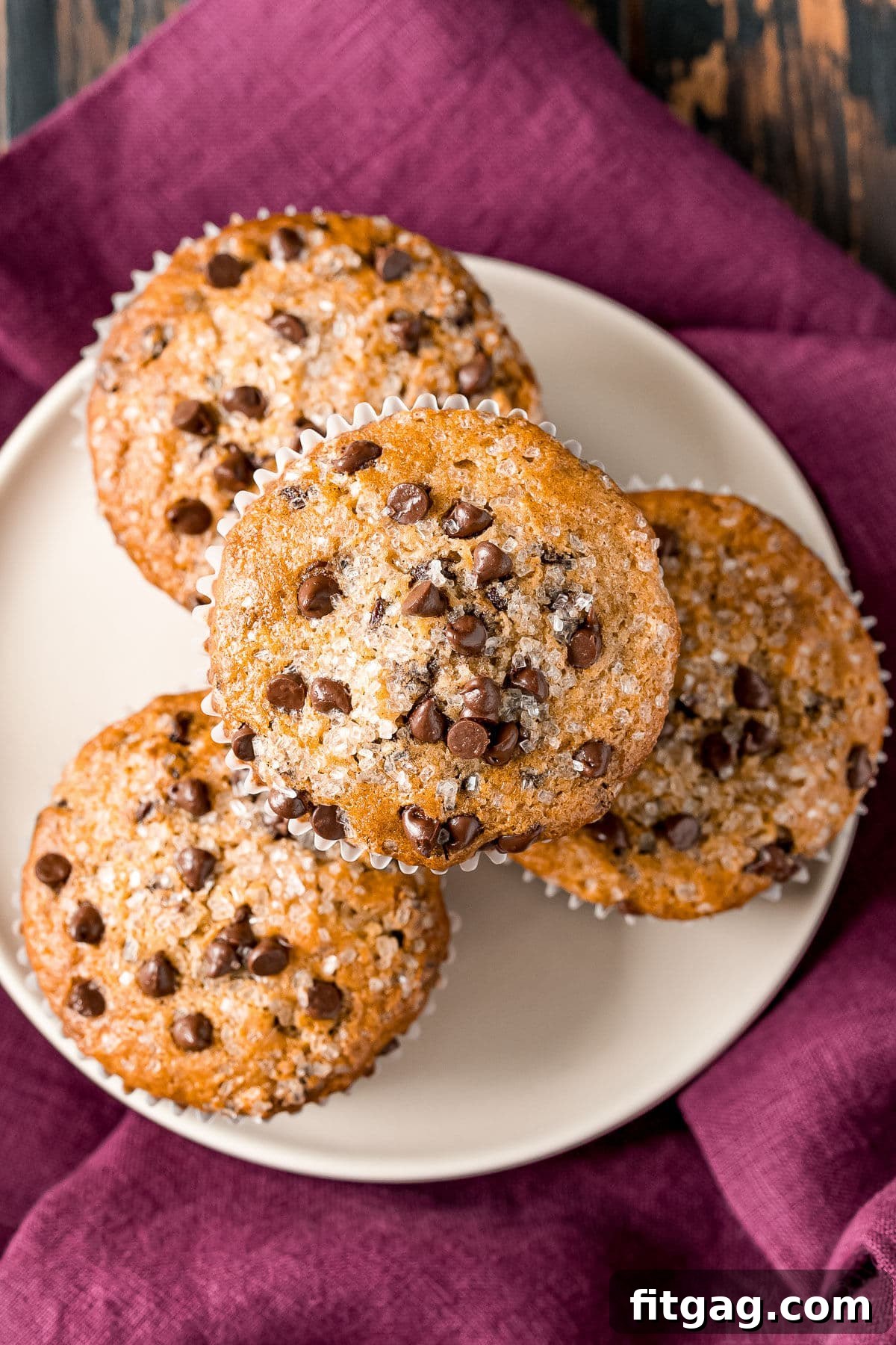 Effortless Chocolate Chip Muffins 10 A beautiful top-down view of chocolate chip muffins artfully arranged on a white plate, resting on a burgundy napkin.