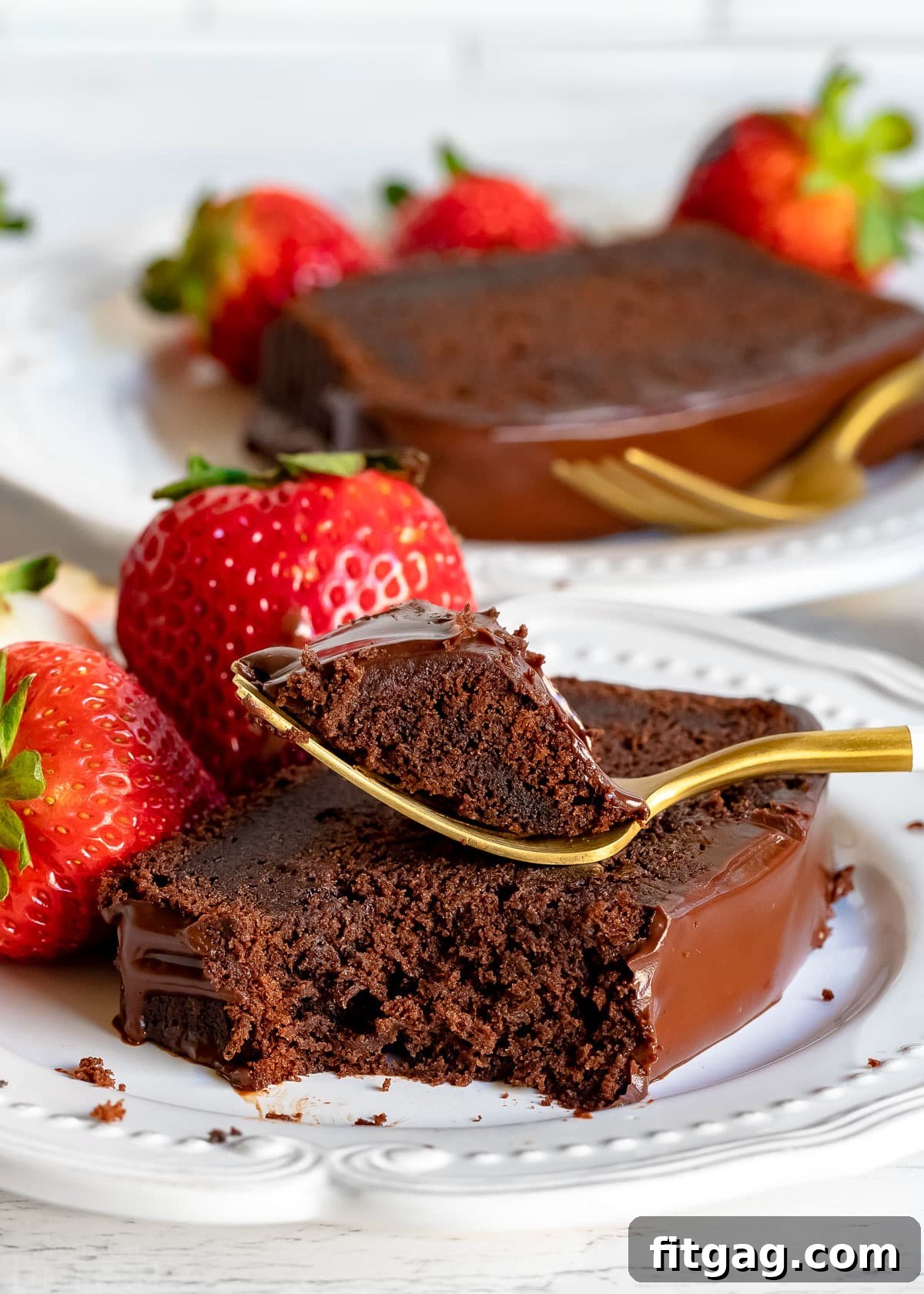 A delectable slice of moist chocolate pound cake, perfectly fudgy, adorned with a luscious chocolate ganache glaze, served on a pristine white plate with a fork, accompanied by fresh strawberries. Another slice of cake and strawberries are visible in the background, enticing chocolate lovers everywhere.