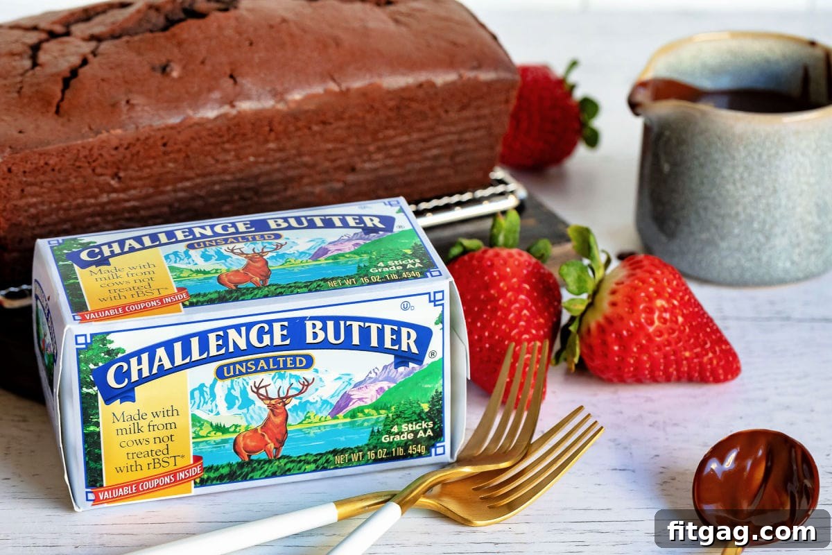 A tempting chocolate pound cake resting next to a box of Challenge Butter, surrounded by a scattering of fresh strawberries and elegant gold forks, emphasizing the quality ingredients used.
