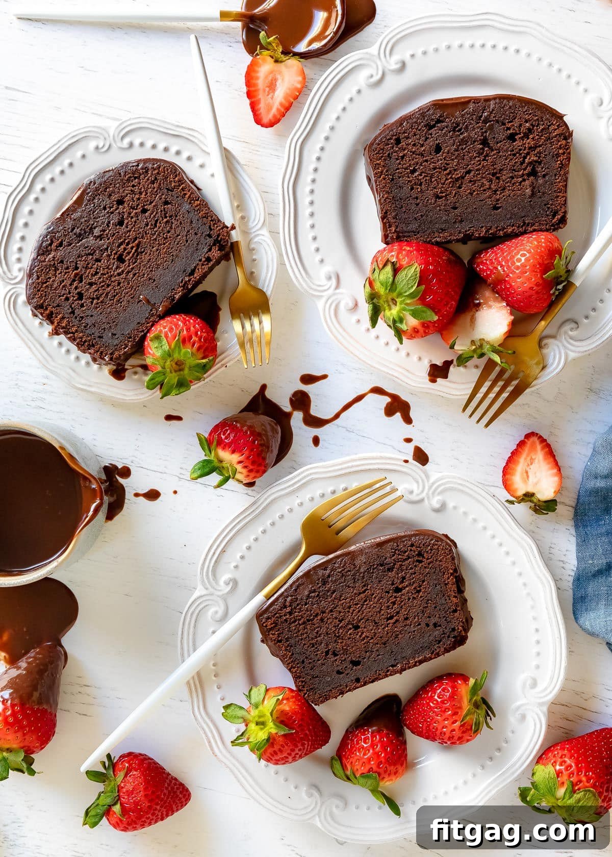A top-down view displaying three perfectly cut slices of chocolate pound cake, elegantly arranged on white plates, each adorned with fresh strawberries and delicate white and gold forks, ready to be enjoyed.