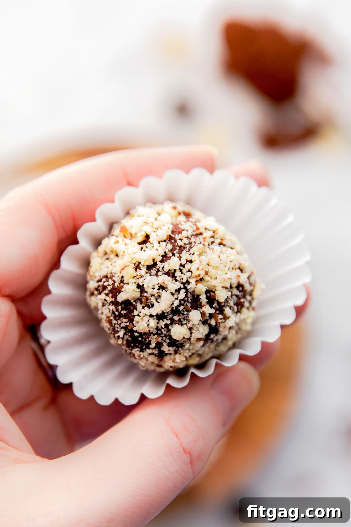 A hand gently holds a single Amaretto chocolate truffle, perfectly coated in finely minced almonds, resting in a pristine white paper liner against a softly blurred wooden backdrop.
