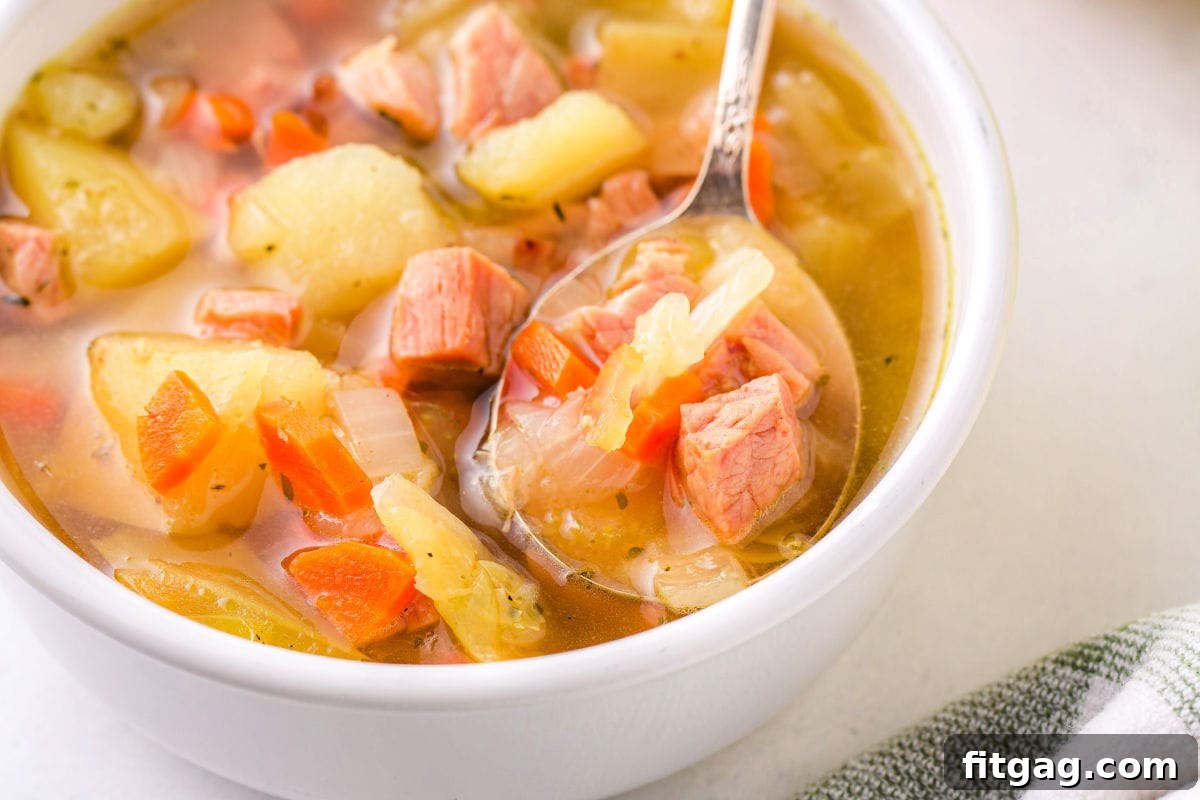 A spoon resting in a white bowl filled with hearty cabbage and ham soup, indicating it's ready to eat.