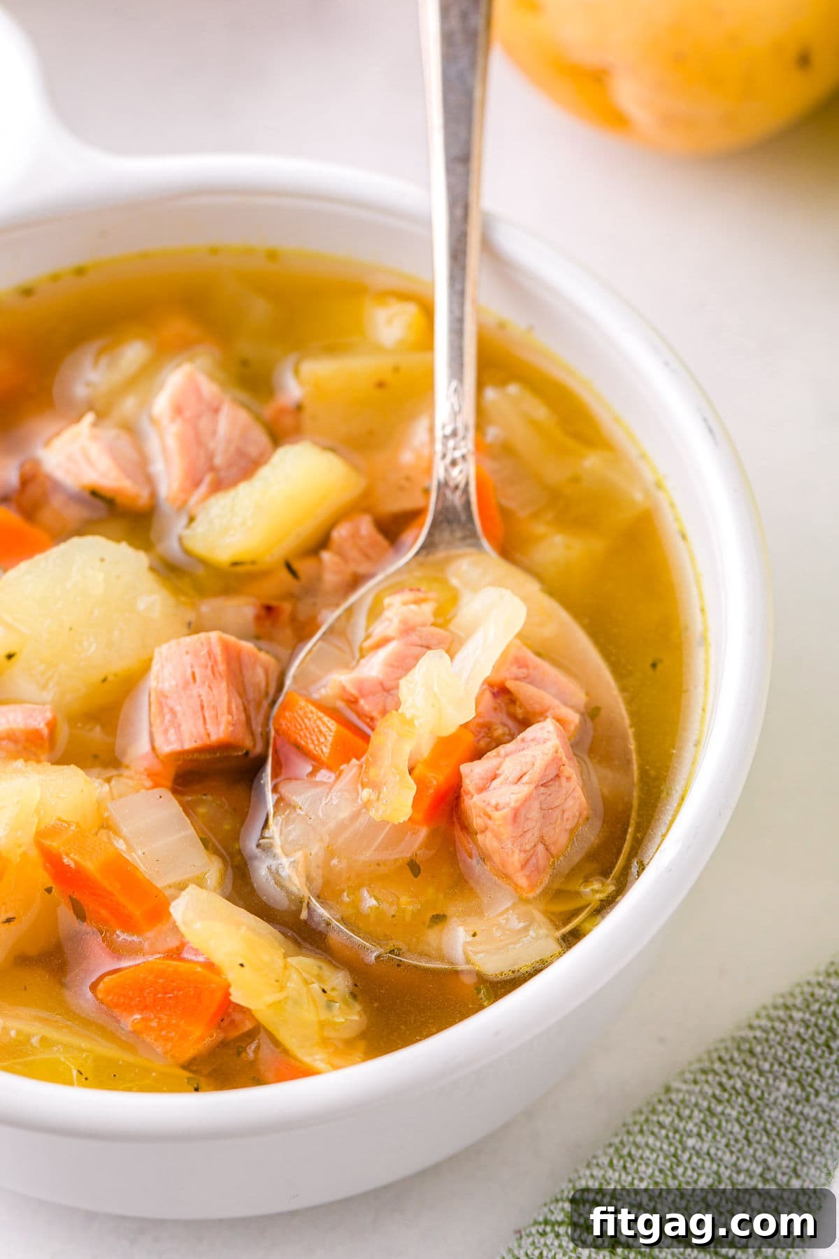 A close-up shot of a spoonful of ham and cabbage soup held above a white bowl, showcasing its rich texture and ingredients.