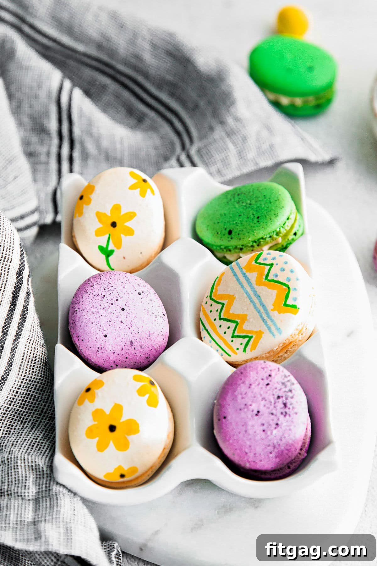 6 macarons decorated like Easter eggs in a white ceramic egg crate.