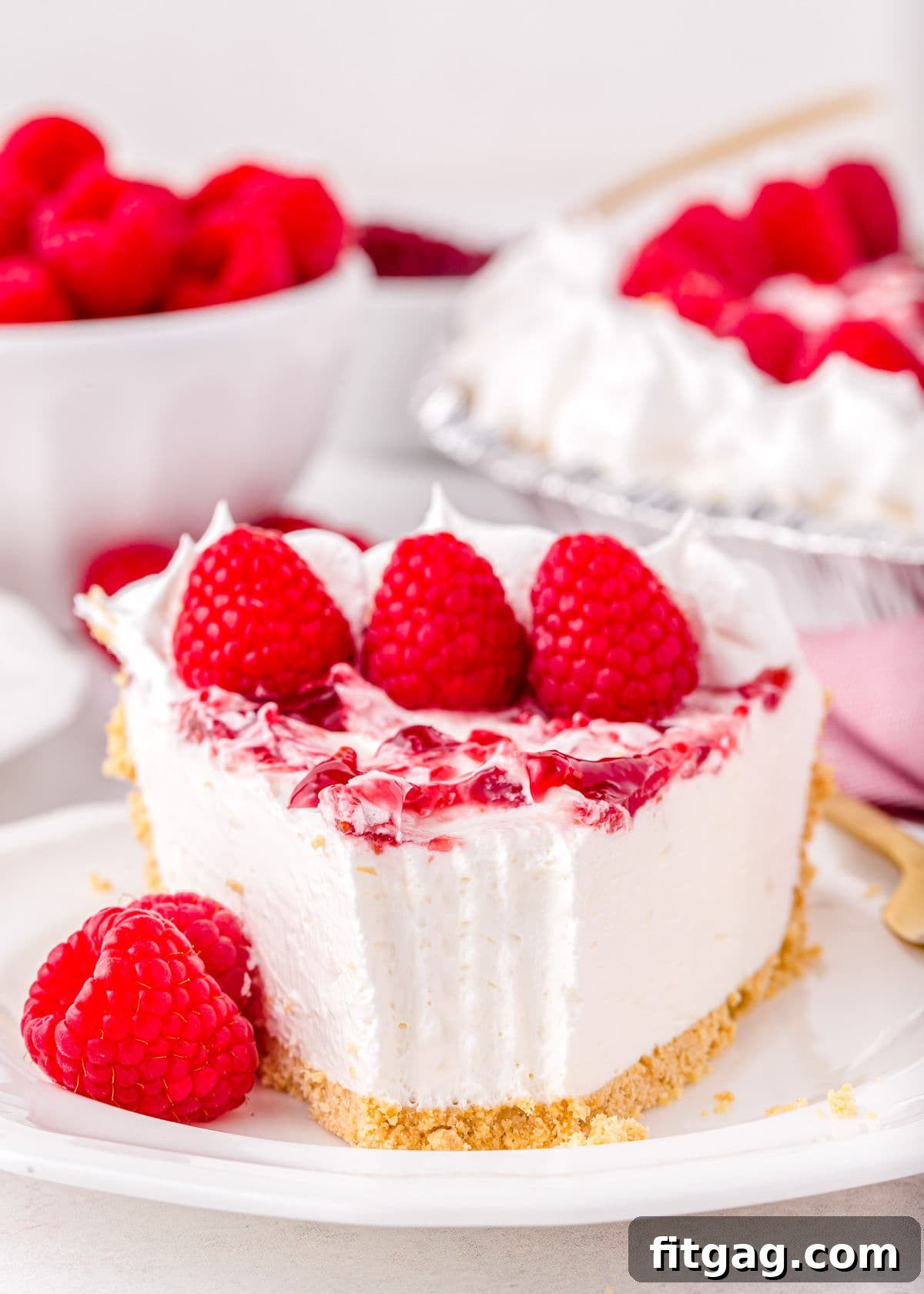 A close-up of a piece of raspberry no-bake cheesecake with a bite taken out, resting on a white plate and garnished with fresh raspberries.