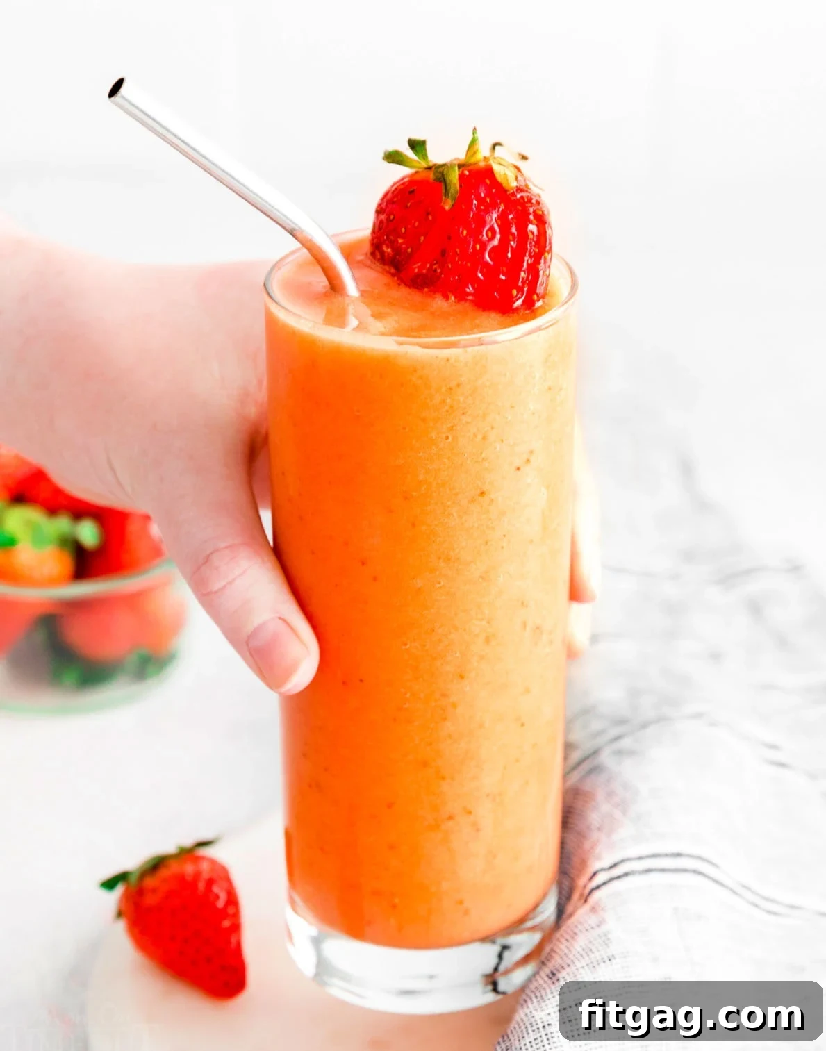 A hand reaching to pick up a tall glass filled with a creamy strawberry mango smoothie, adorned with a fresh strawberry slice on the rim.