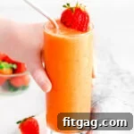 A hand picking up a glass of strawberry mango smoothie, garnished with a sliced strawberry.