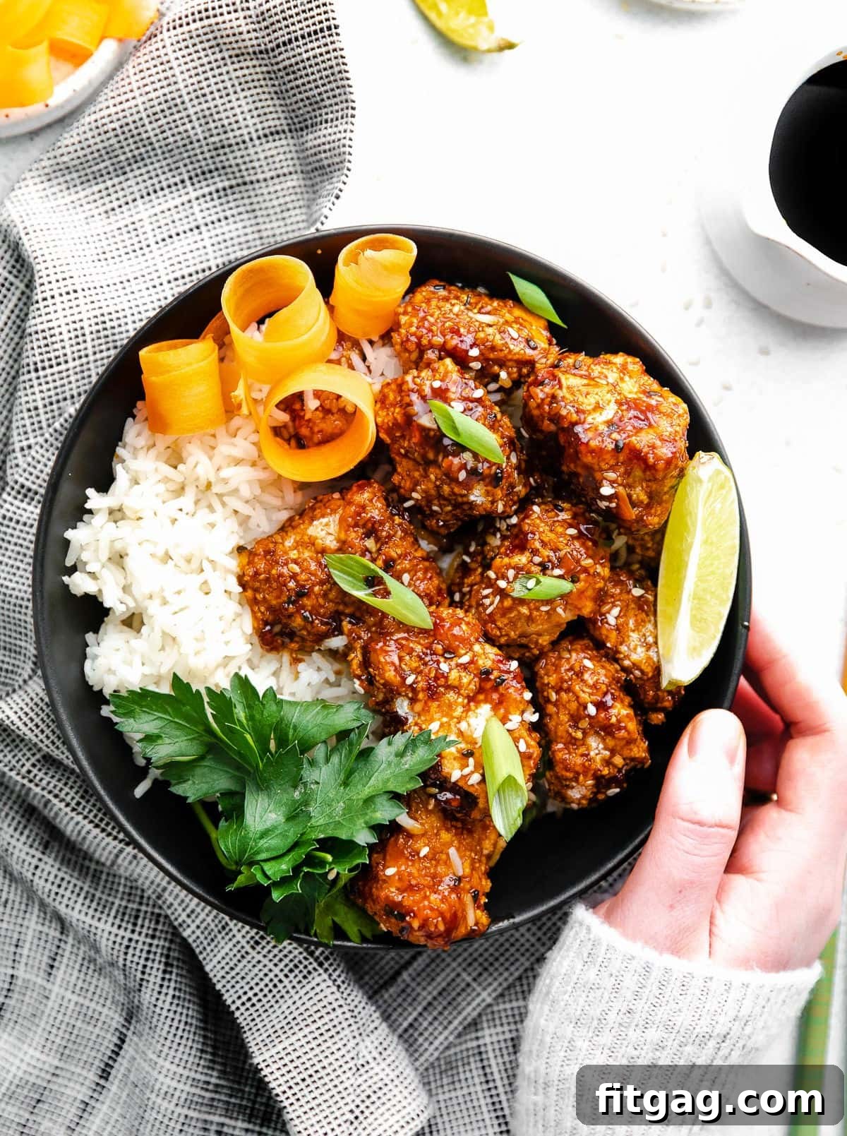 Black bowl with rice and teriyaki chicken garnished with orange peels, lime wedge and parsley.