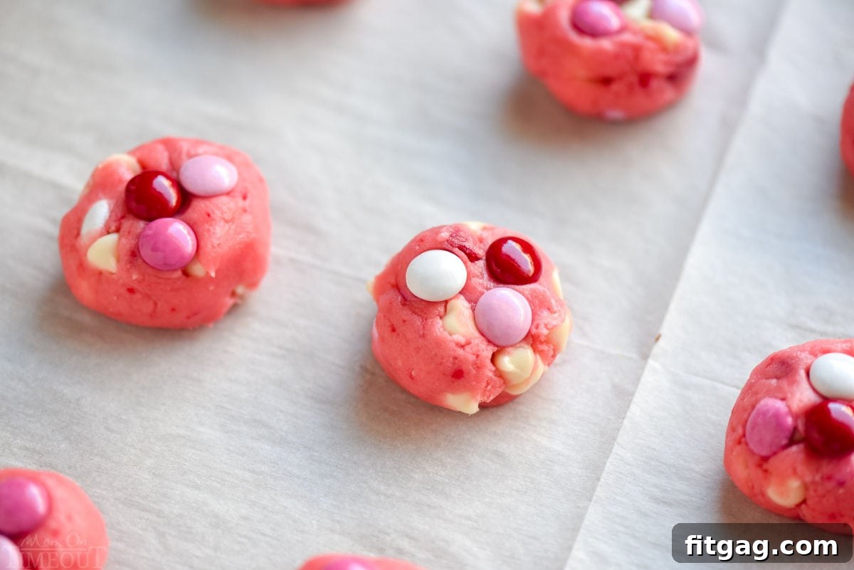Perfectly portioned cookie dough balls, generously topped with M&Ms, arranged on a parchment-lined baking sheet and ready to be transformed into delicious cookies.