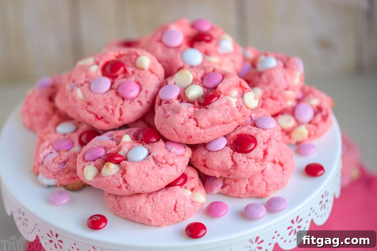 A beautiful display of Strawberry Cake Mix Cookies, generously studded with M&Ms and white chocolate chips, stacked high on a elegant white metal cake stand.