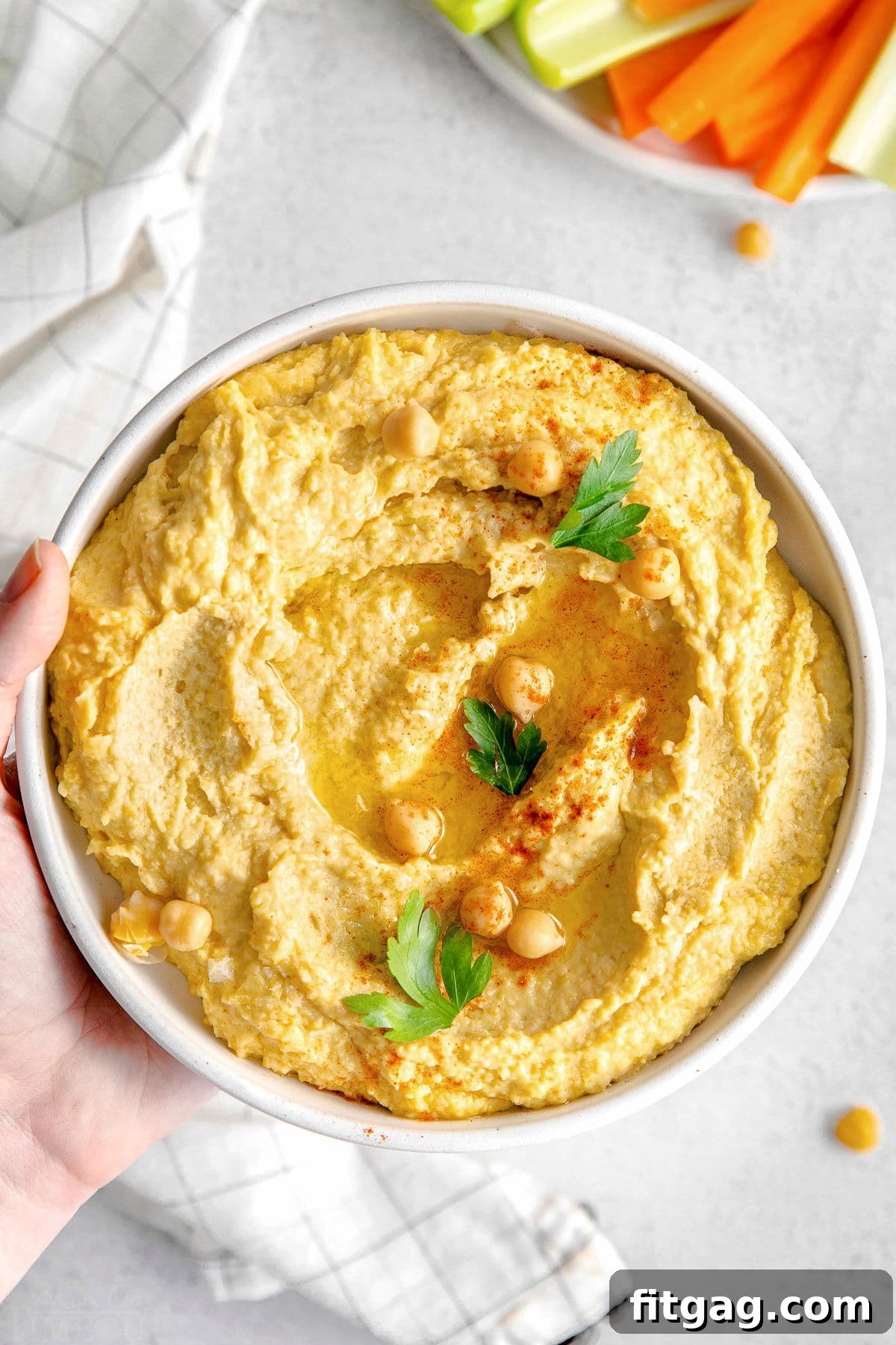 A hand holds a small white bowl filled with creamy hummus, invitingly garnished with olive oil and a sprinkle of paprika, ready to be enjoyed.
