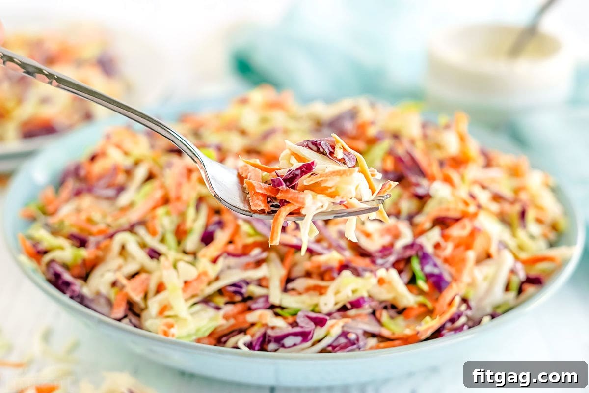 A forkful of creamy coleslaw held against the backdrop of a large bowl filled with the fresh salad.