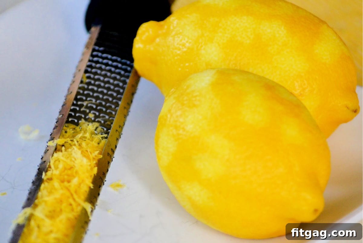 Close-up image of a hand zesting a fresh lemon over a white plate with a microplane, showcasing the process of collecting lemon zest.