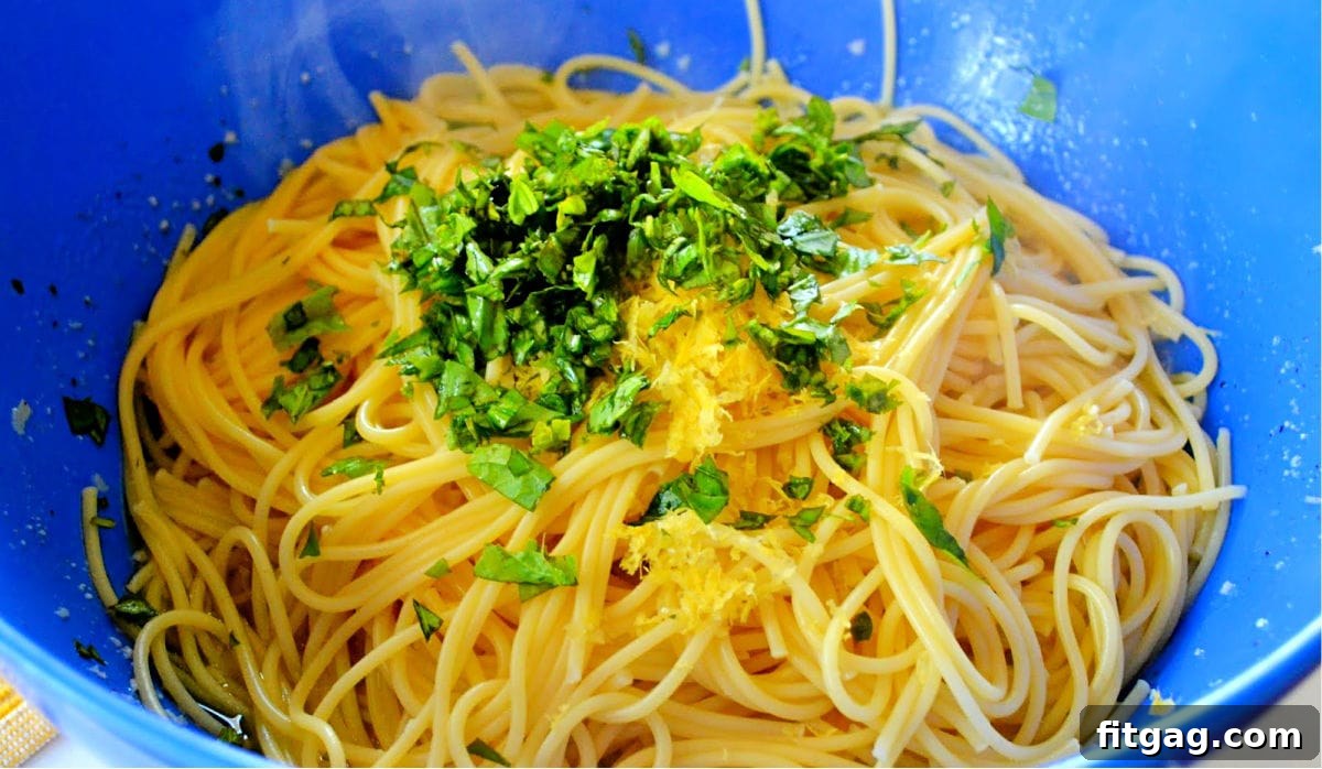 A large blue mixing bowl filled with freshly cooked spaghetti, lemon sauce, basil, and lemon zest, being tossed together.