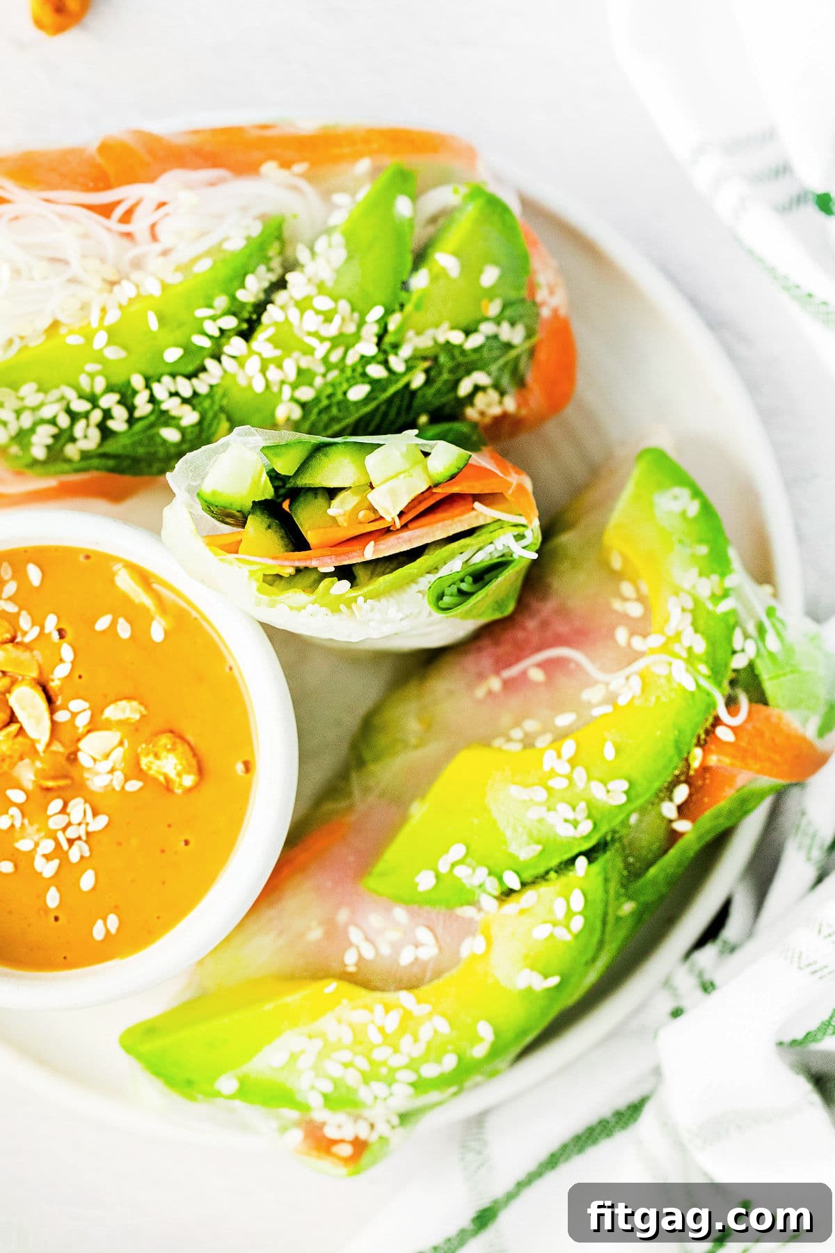 Top-down view of summer rolls on a white plate with one roll cut in half, alongside a small bowl of peanut dipping sauce.