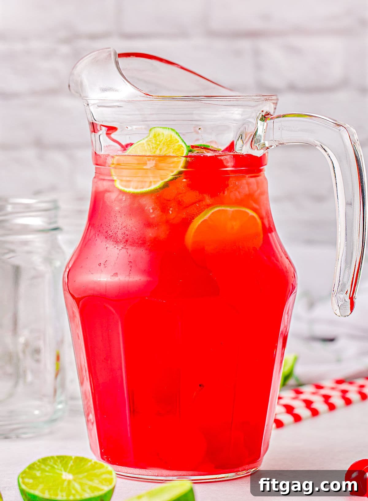 A large, elegant glass pitcher overflowing with vibrant cherry limeade, beautifully adorned with fresh lime slices floating within and surrounded by additional lime wedges on the counter. It's perfectly prepared and ready to be poured and enjoyed.