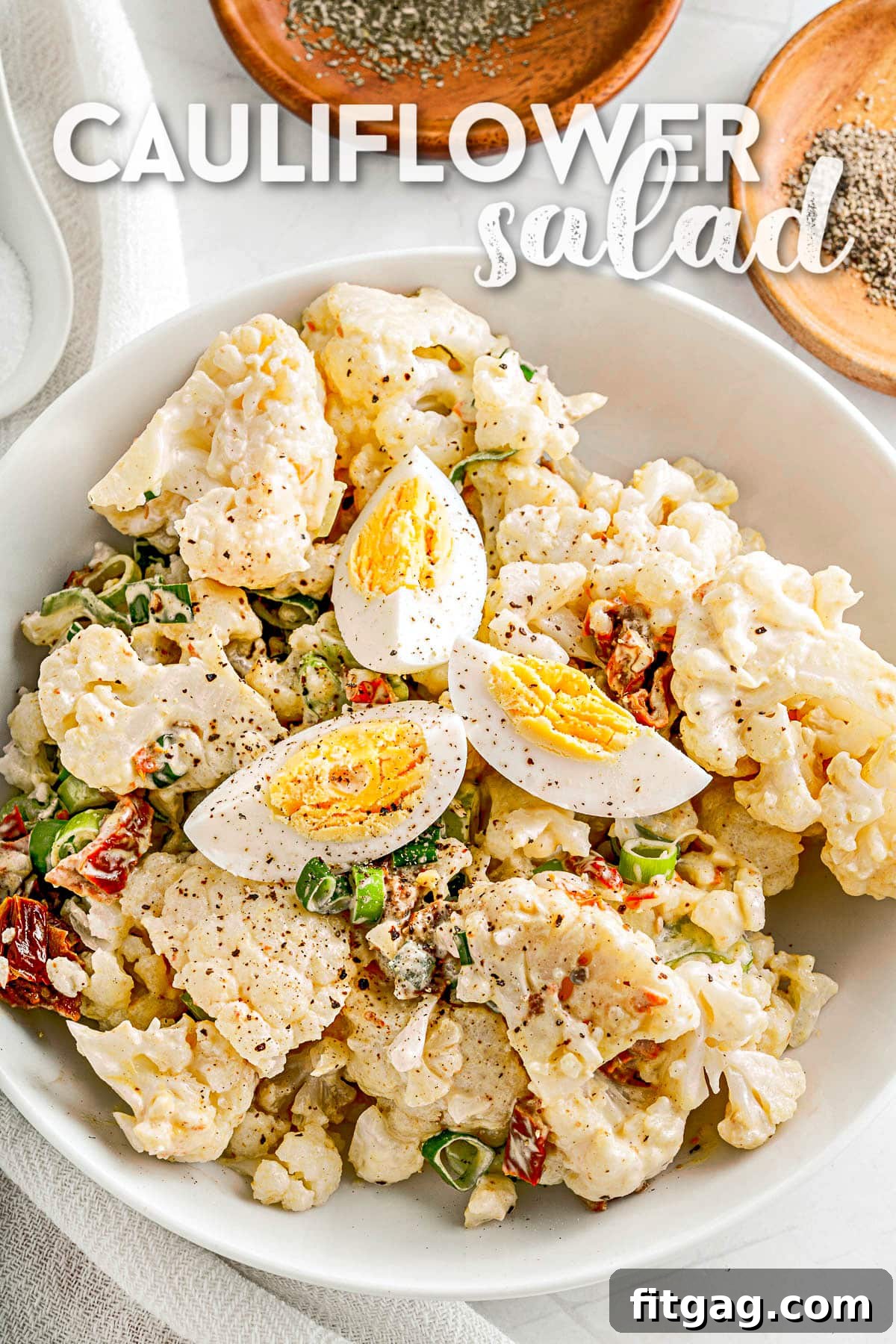 Cauliflower salad made like a potato salad in a white serving bowl, topped with slices of hard-boiled egg and a sprinkle of paprika.
