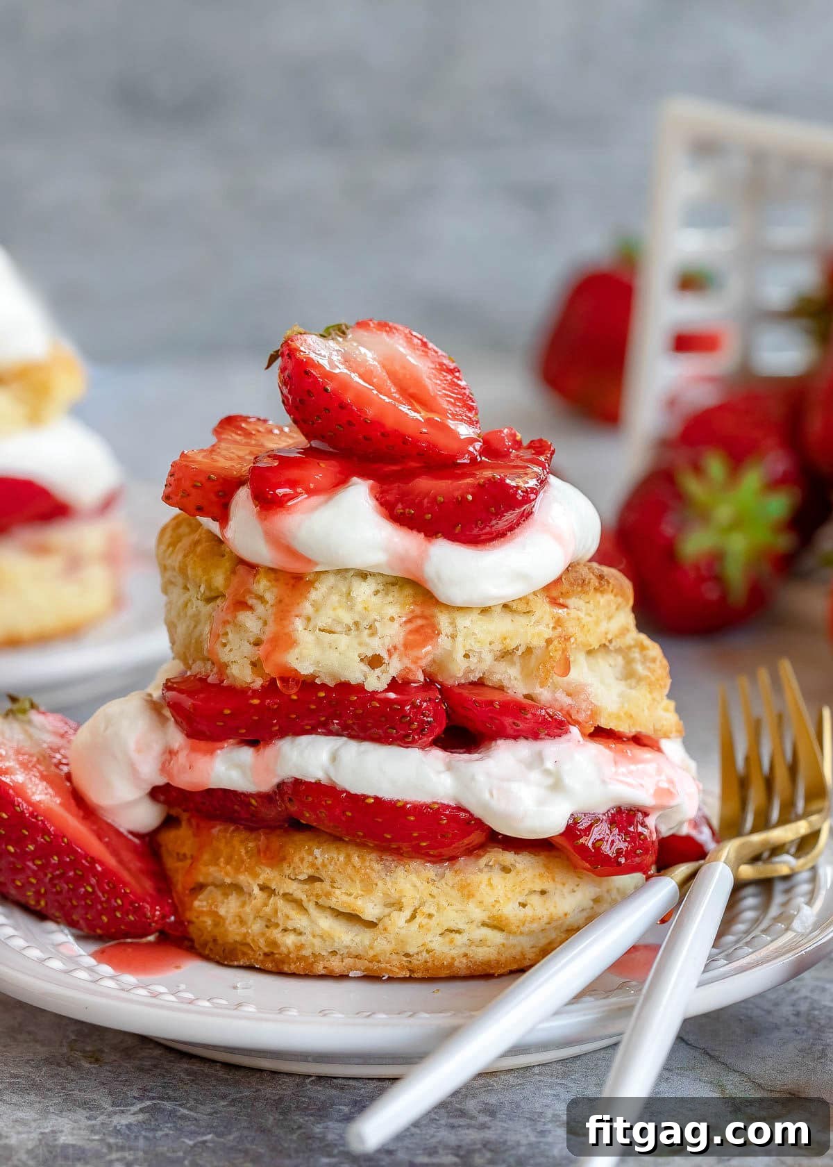 A delectable strawberry shortcake featuring homemade biscuits, perfectly macerated strawberries, and fresh whipped cream, layered artfully on a white plate with two forks ready for enjoyment.