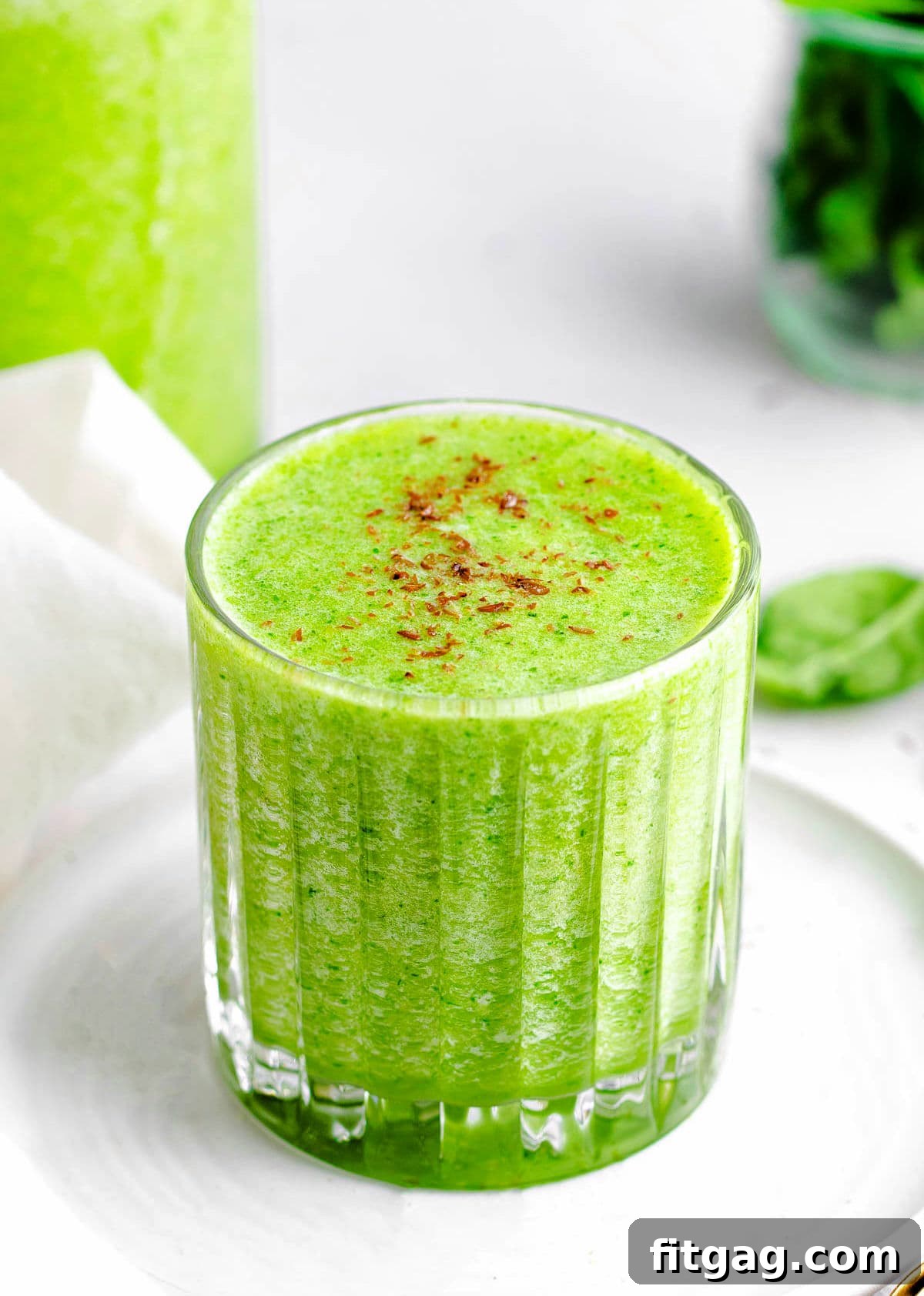 The Perfect Green Smoothie 2 A tall glass of vibrant green smoothie sitting on a small white plate, with a blender and fresh greens blurred in the background, suggesting freshness and homemade goodness.