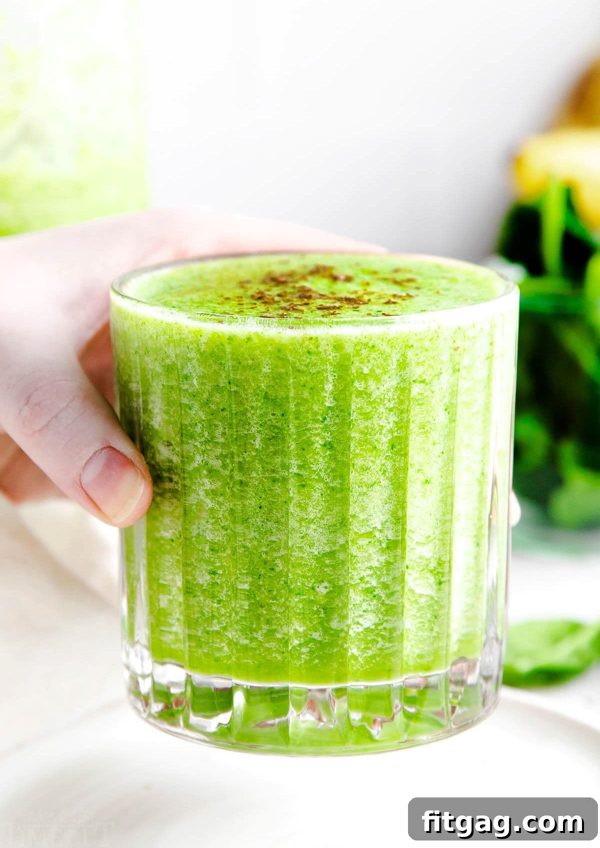 The Perfect Green Smoothie 3 A hand holding a ribbed glass filled with a bright green smoothie, with a lush green, leafy background suggesting natural ingredients and a fresh, healthy lifestyle.