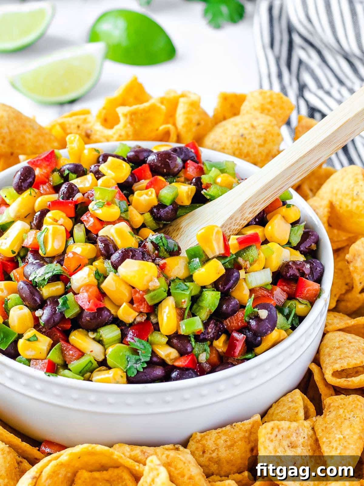 Black bean and corn salsa in a white bowl with a wooden spoon, surrounded by corn chips and fresh limes.