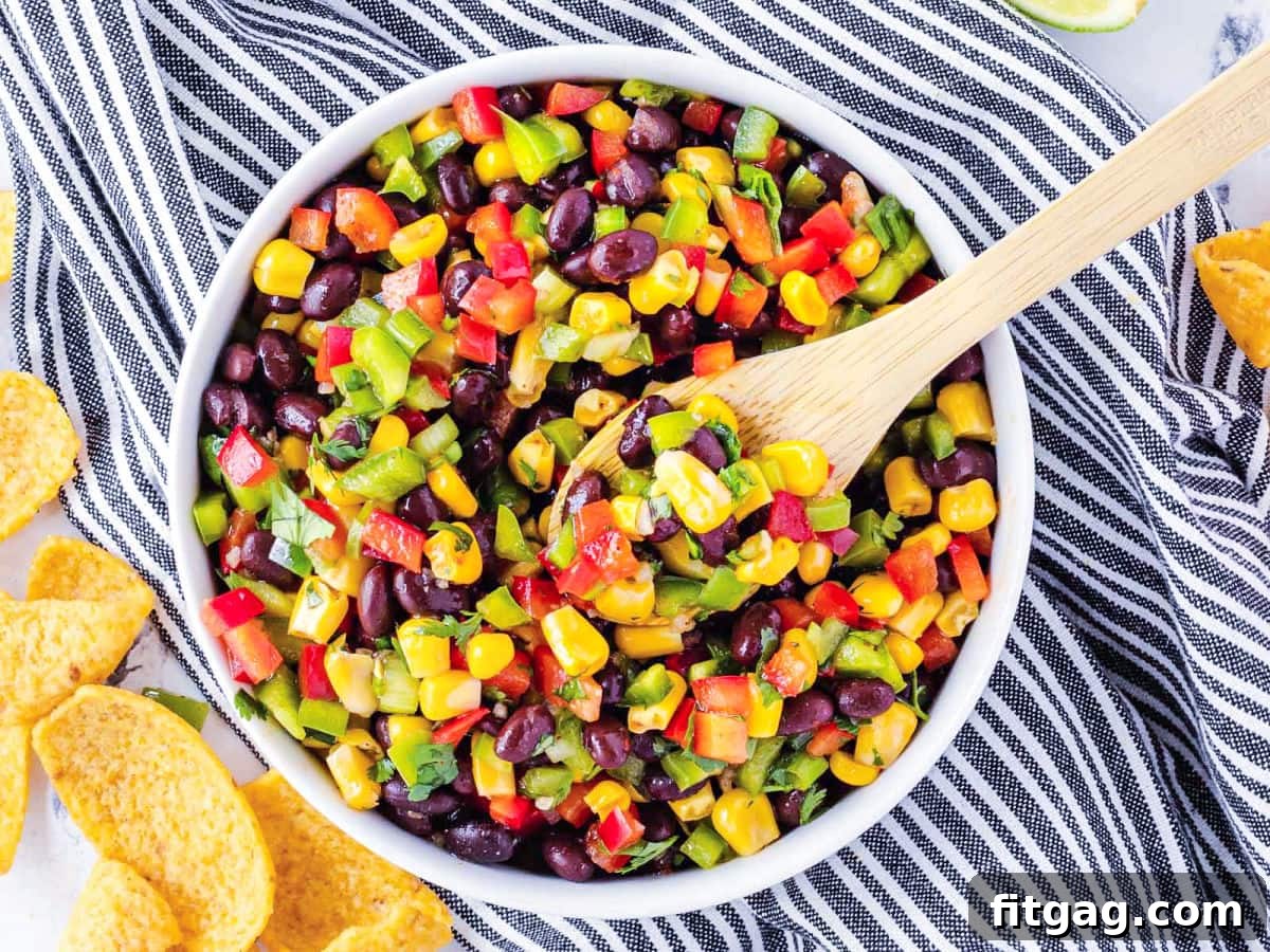 Black bean corn salsa in a white bowl with a wooden serving spoon, placed on a black and white striped towel.