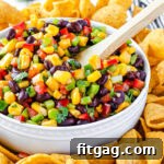 Black bean and corn salsa in a white bowl with a wooden spoon, surrounded by corn chips and fresh limes.