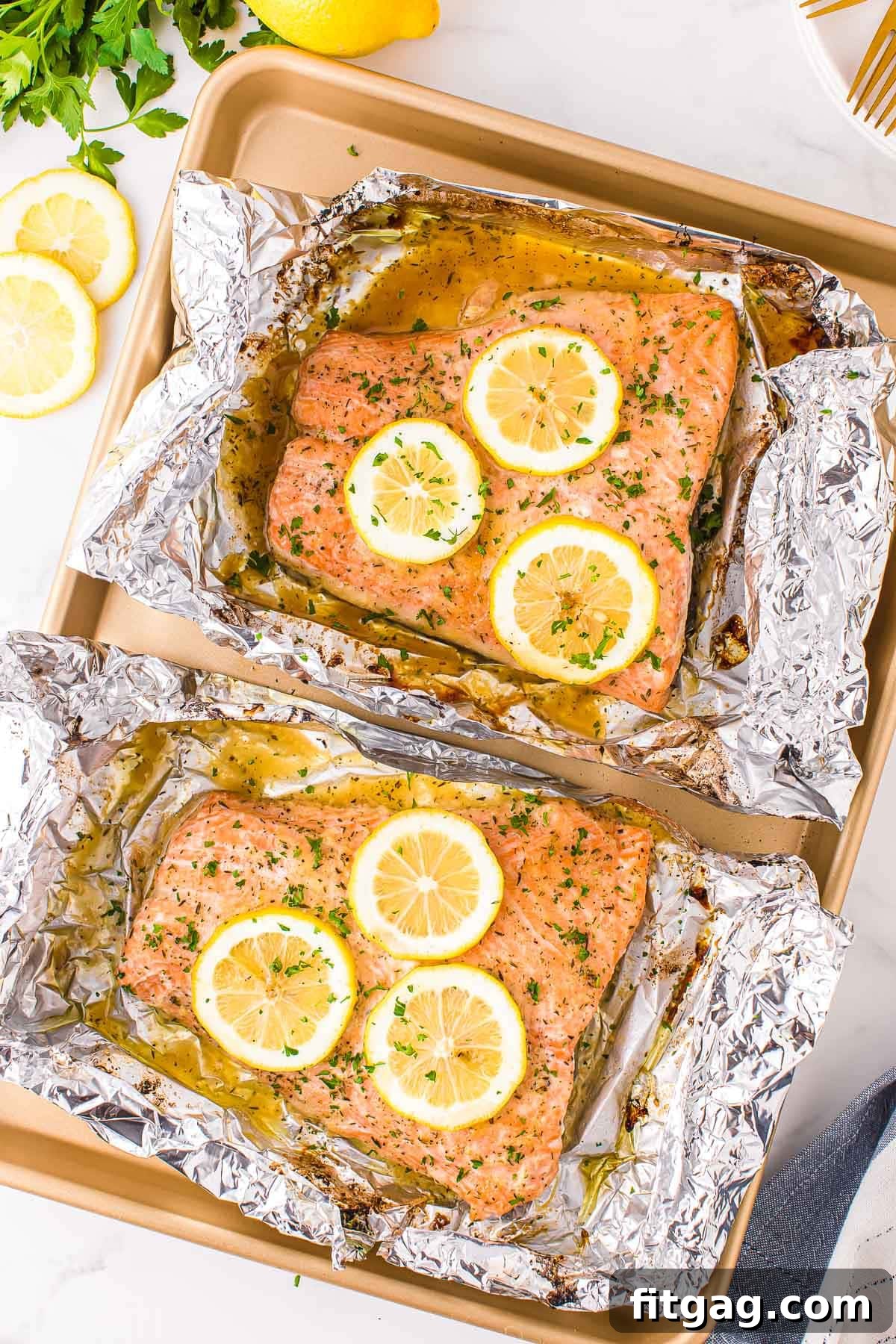 Two large salmon fillets baked in foil, resting on a gold sheet pan, adorned with lemon slices and fresh minced parsley.