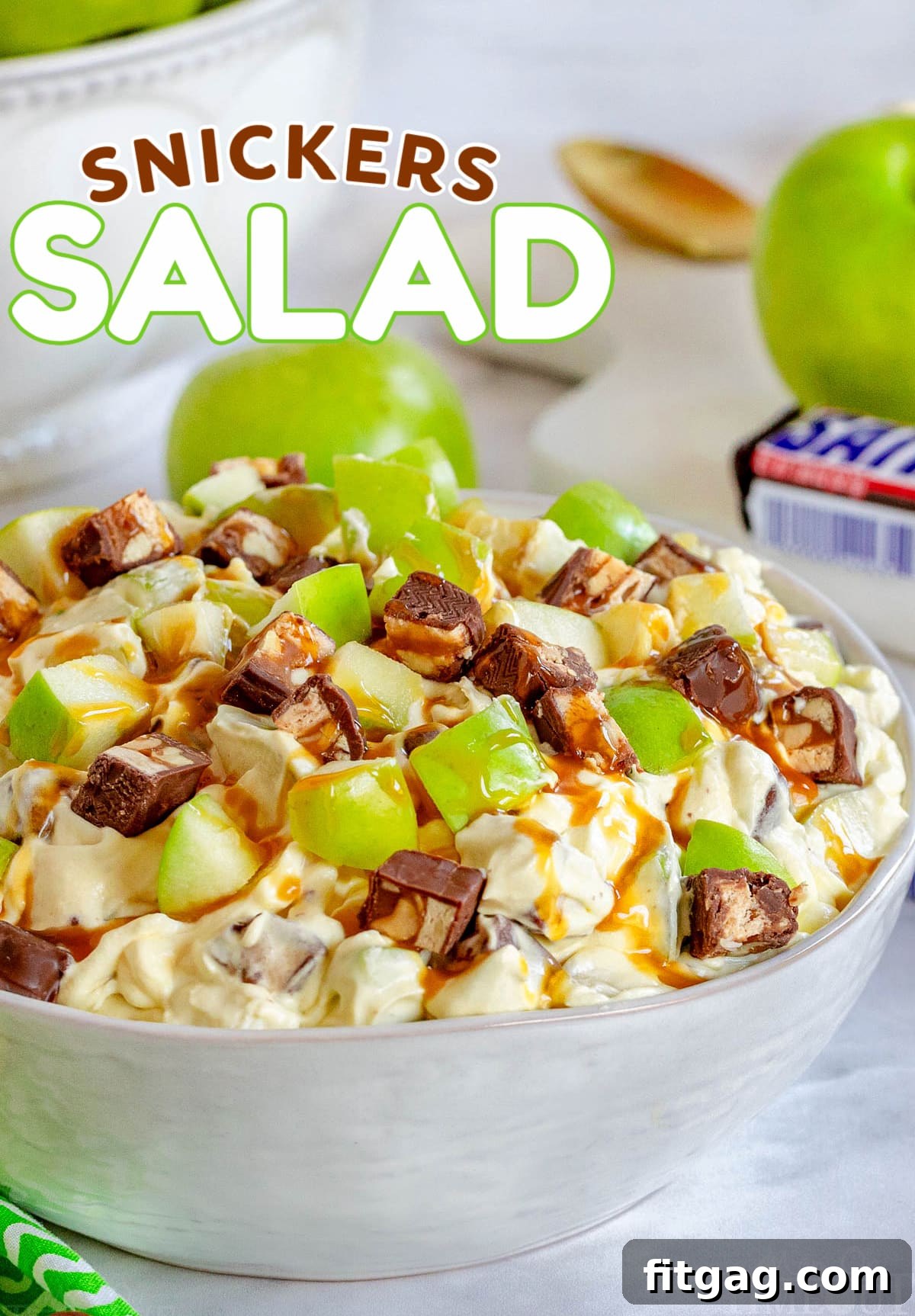 Snickers Caramel Apple Delight 2 snickers salad in white salad bowl topped with a drizzle of caramel sauce. green apples and snickers candy bar in background. title overlay at top of image.