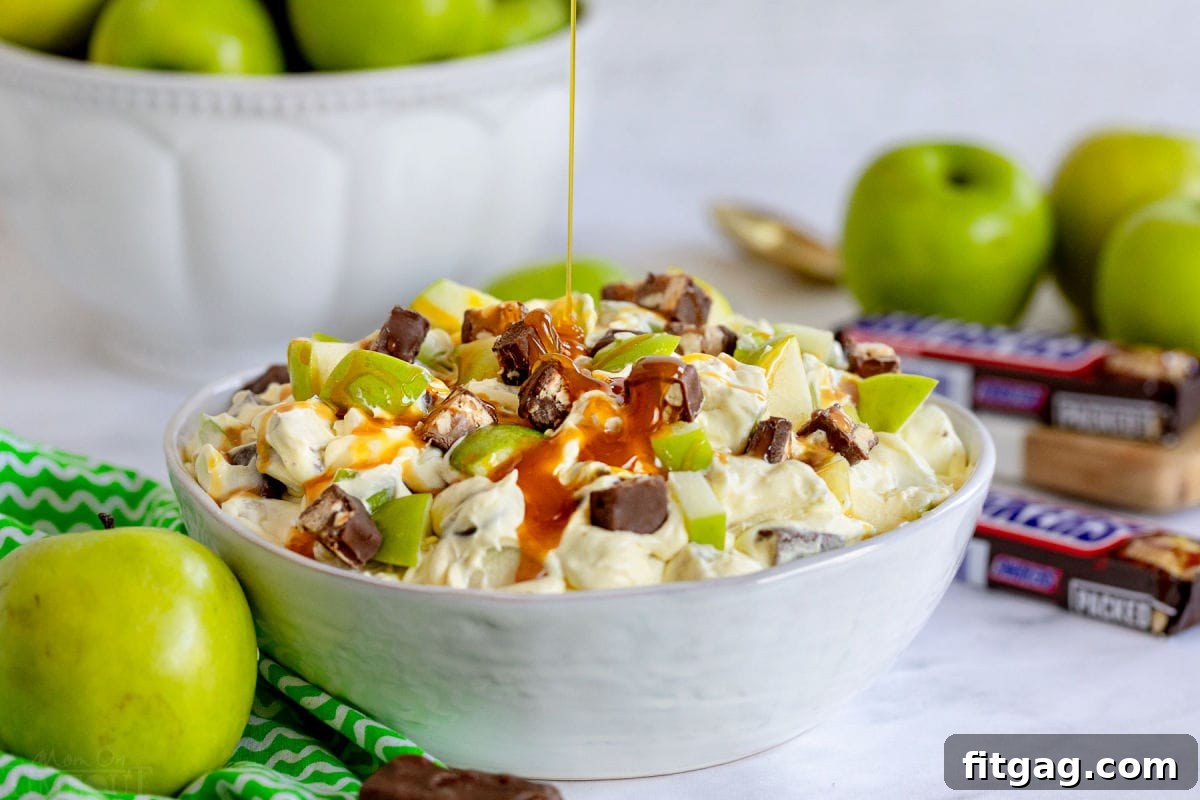 Snickers Caramel Apple Delight 7 caramel sauce being drizzled over the top of the snickers salad in large white bowl. apples and snickers around the bowl.