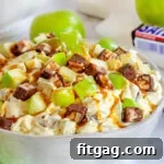 snickers salad in white salad bowl topped with a drizzle of caramel sauce. green apples and snickers candy bar in background.