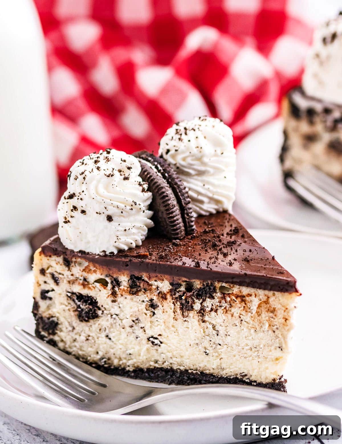 Ultimate Oreo Cheesecake 3 A perfectly cut slice of Oreo cheesecake rests on a white plate, with a rustic red checkered napkin softly blurred in the background, highlighting the dessert's readiness to be enjoyed.