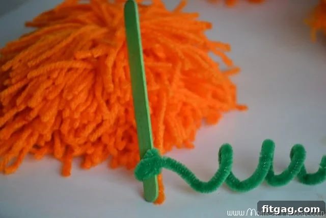 A popsicle stick stem with a green pipe cleaner curled around it, ready to be attached to a pom pom pumpkin.