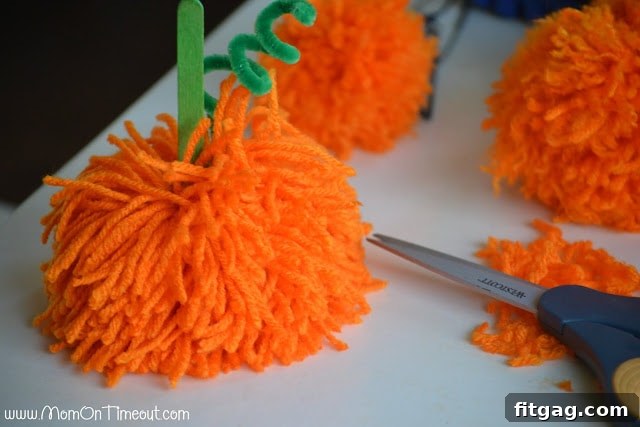 A person inserting a decorated popsicle stick stem into the center of an orange pom pom, preparing to glue it in place for a DIY pumpkin.
