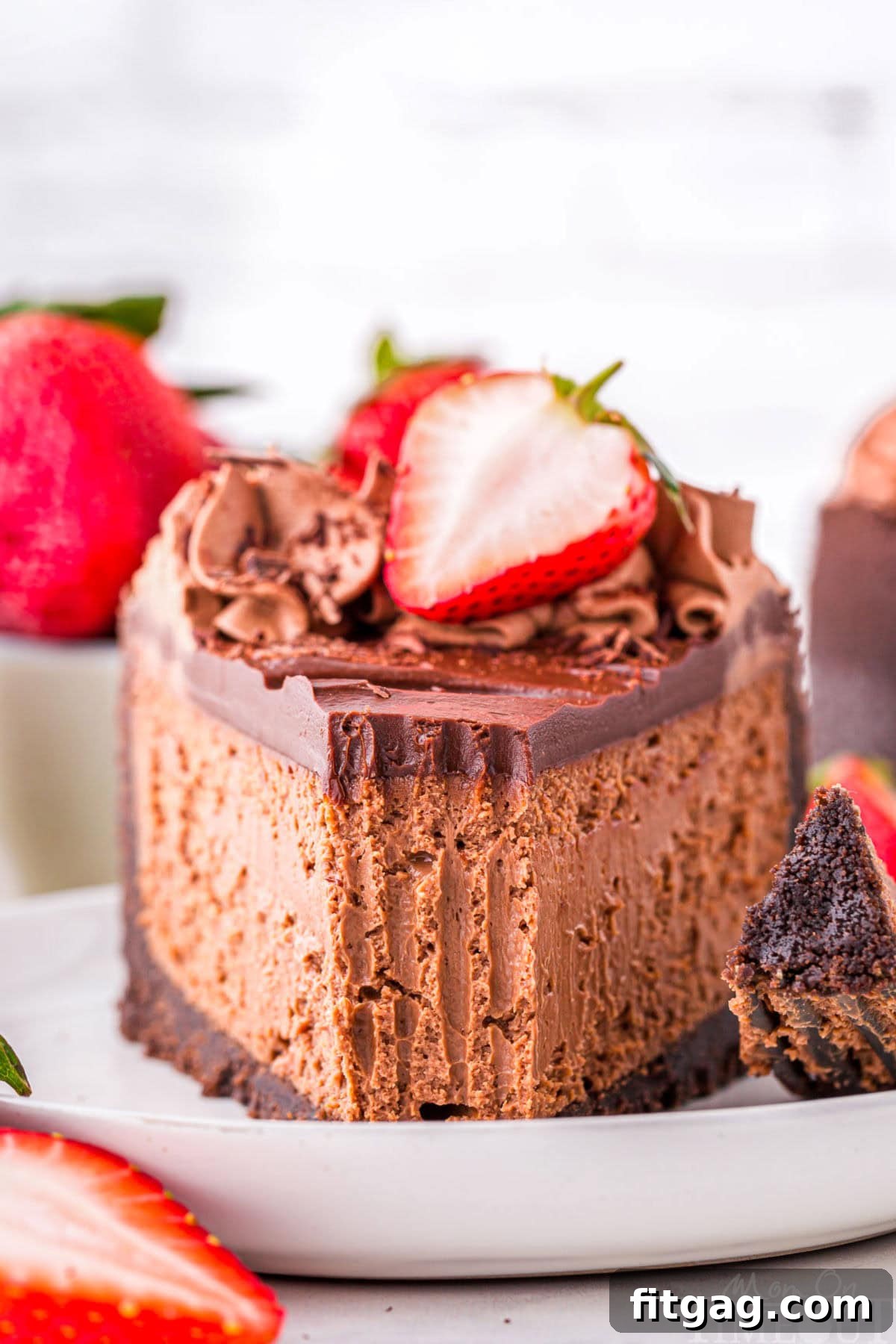 Slice of decadent chocolate cheesecake with an oreo crust and rich chocolate ganache on white round plate. Topped with chocolate shavings and fresh strawberries.