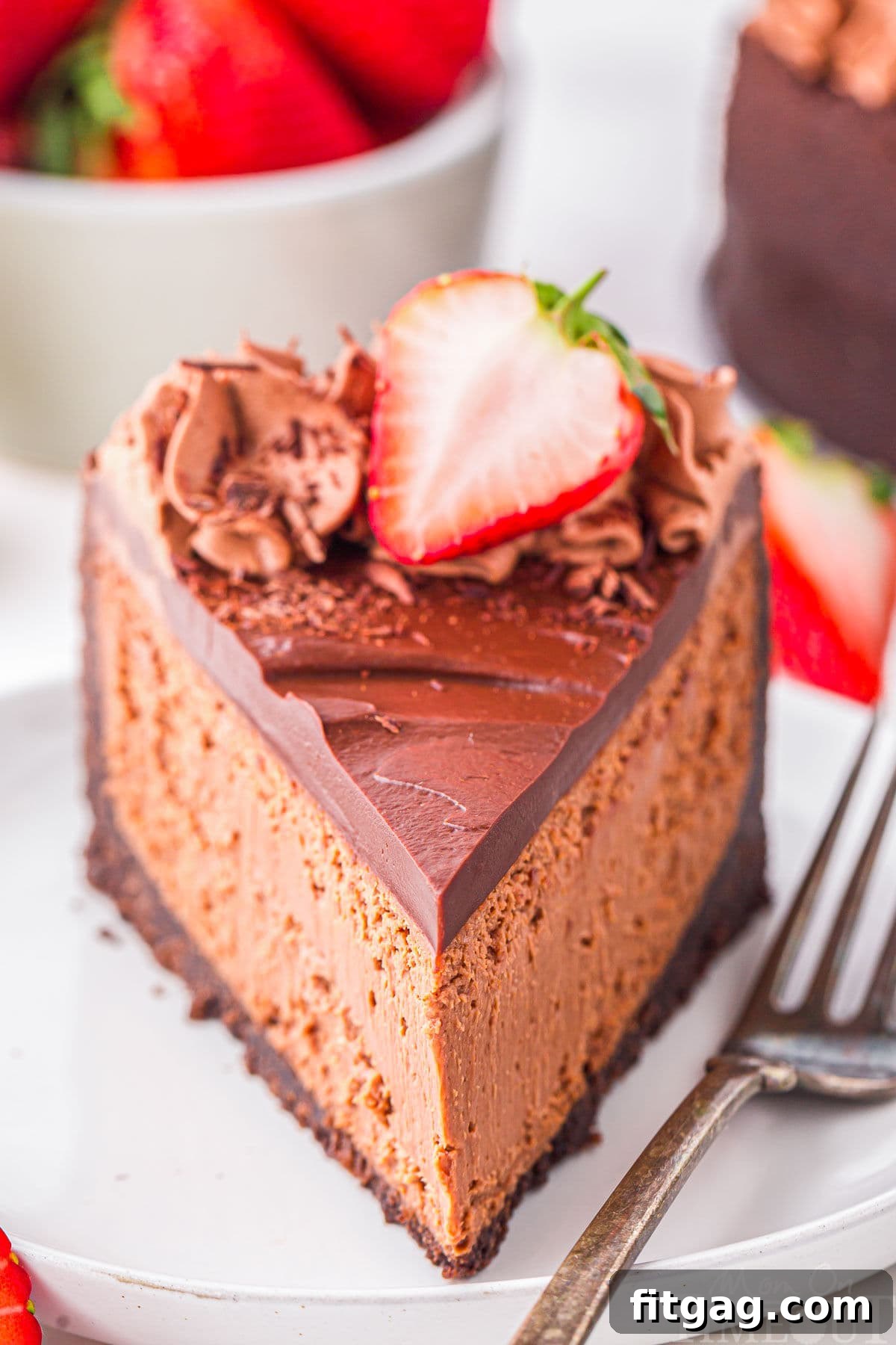 Slice of decadent chocolate cheesecake with an oreo crust and rich chocolate ganache on white round plate. Topped with chocolate shavings and fresh strawberries. Strawberries and more cheesecake can be seen in the background.