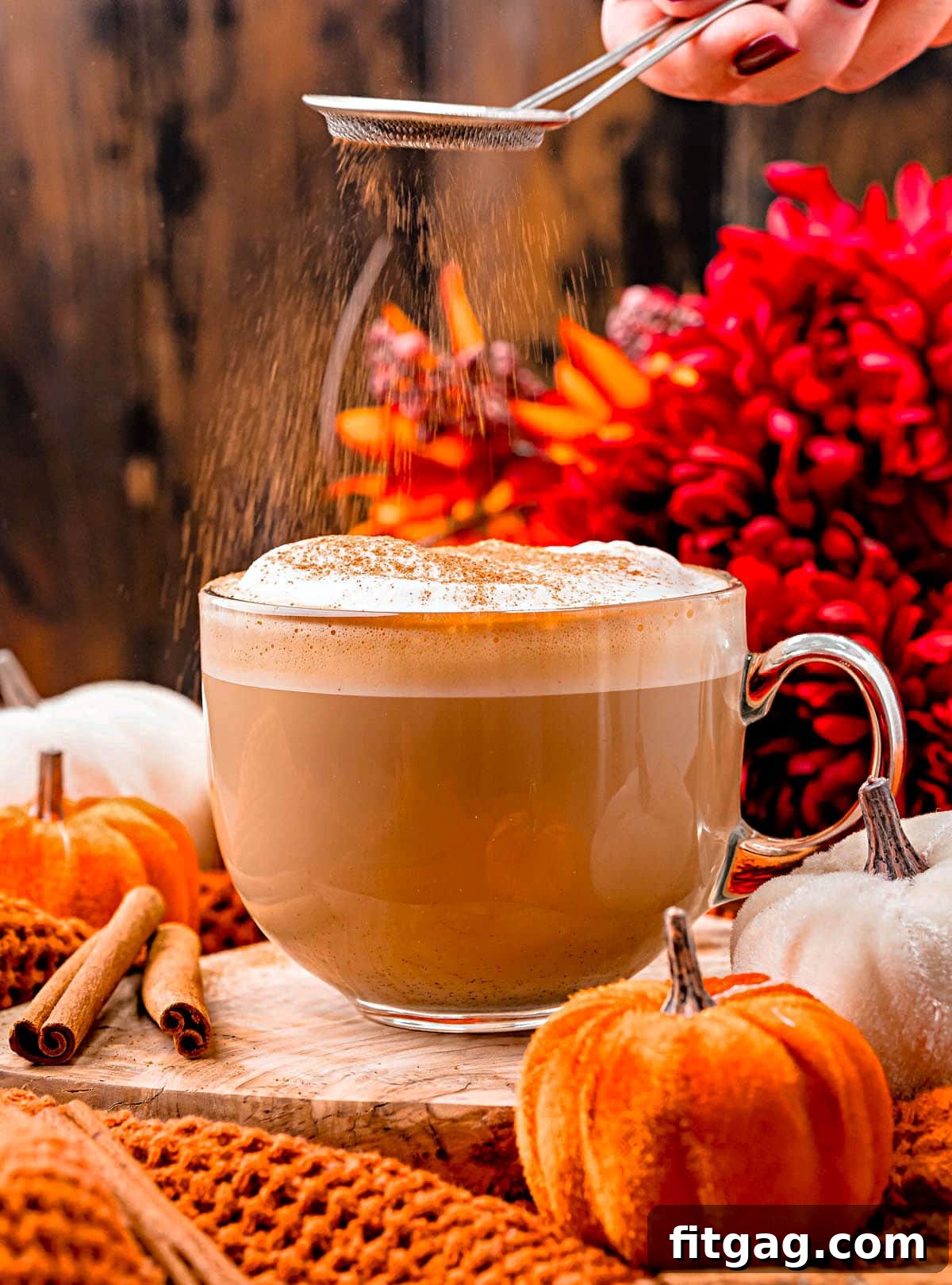 pumpkin spice latte in glass mug with pumpkin spice being dusted on top of the foam topping. colorful fall flowers in background and small velvet pumpkins scattered about.