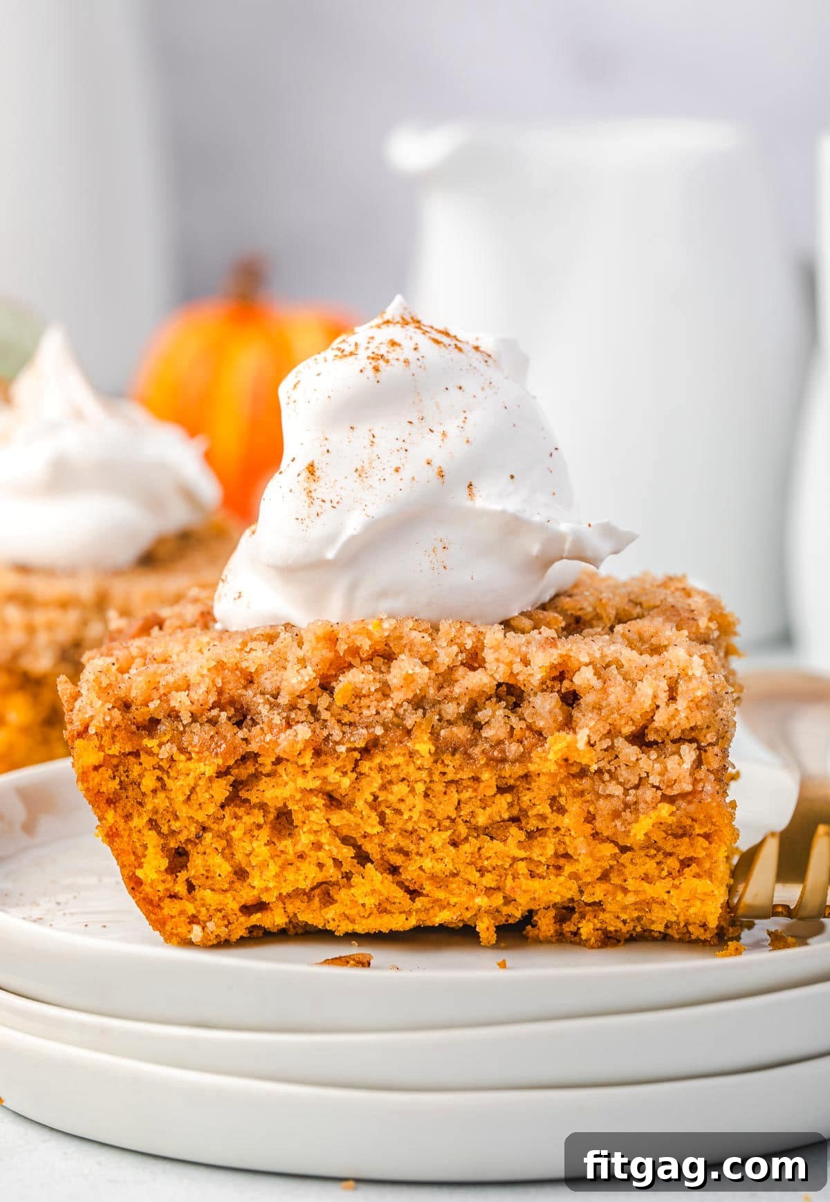 A delectable slice of Pumpkin Coffee Cake, beautifully presented on a stack of white plates and generously topped with fluffy whipped cream, highlighting its warm, inviting colors and inviting texture.