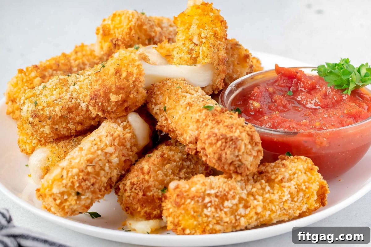 Golden Gooey Air Fryer Mozzarella Sticks 3 A close-up view of numerous golden-brown mozzarella sticks, freshly cooked in an air fryer, resting on a white plate next to a small dish of rich marinara dipping sauce.