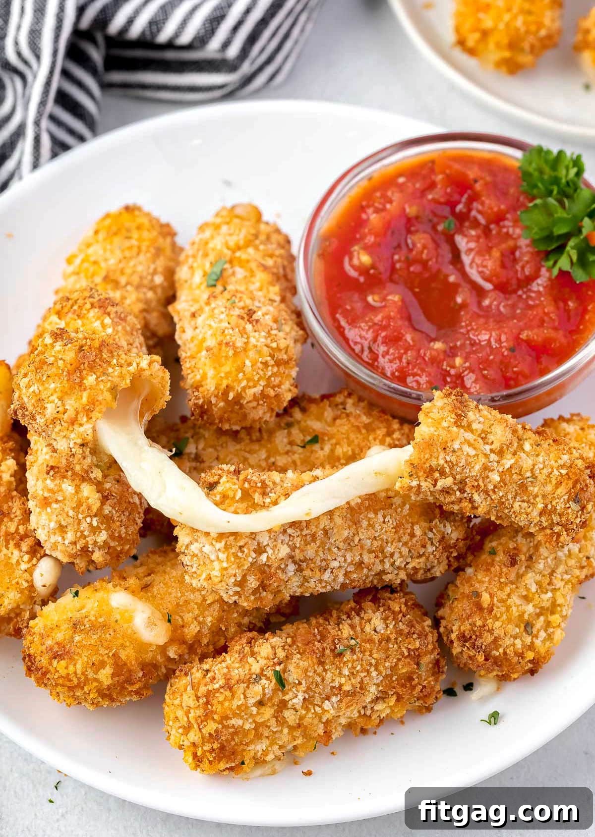 Golden Gooey Air Fryer Mozzarella Sticks 7 A delectable cheese stick with an impressive cheese pull, resting atop a mound of other perfectly air-fried mozzarella sticks on a pristine white plate, showcasing the gooey interior.