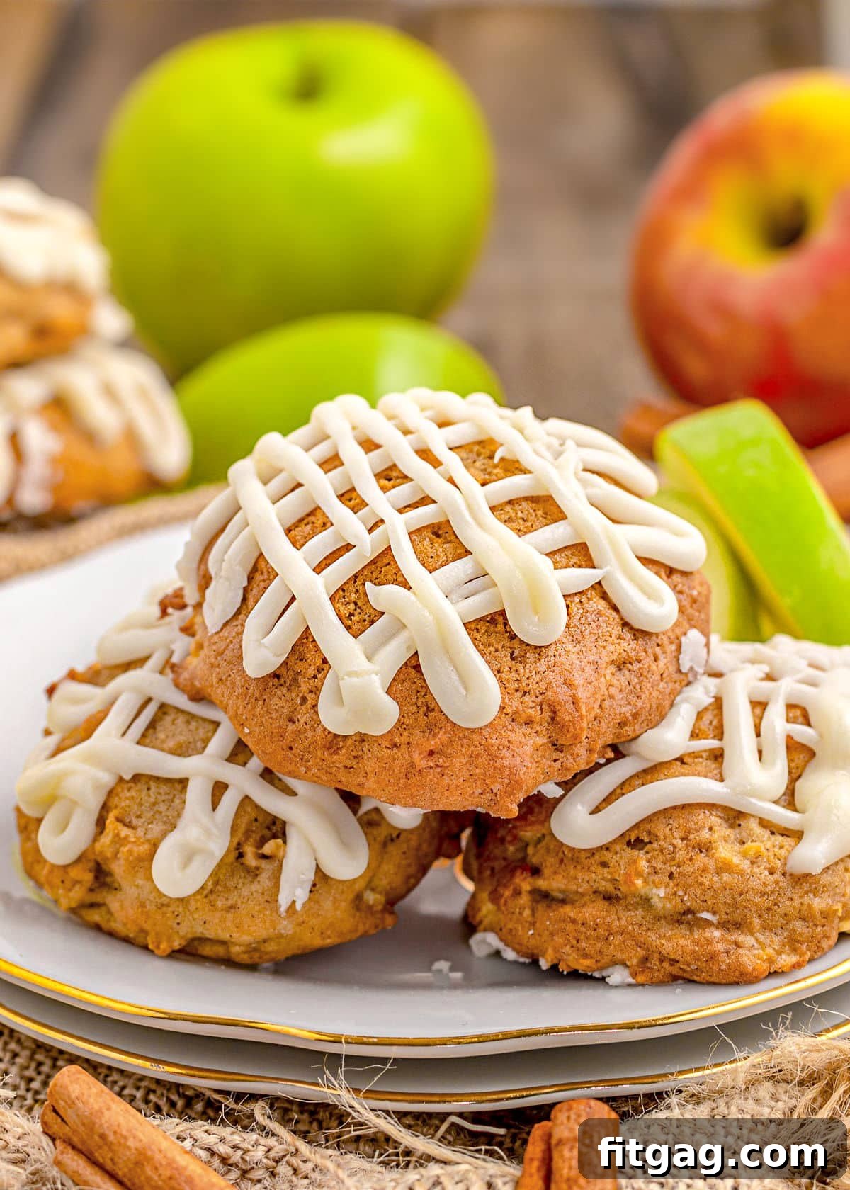 Three freshly baked apple cookies with a generous drizzle of maple icing, resting on a white plate with a delicate gold rim. Fresh whole apples and sliced apples are artfully arranged in the background, hinting at the key ingredient.