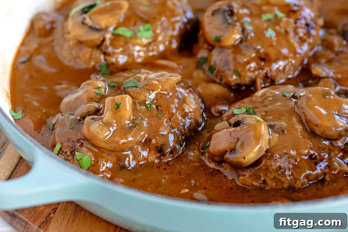 Easy Homestyle Salisbury Steak 4 A close-up view of three Salisbury steaks glistening with mushroom gravy in a deep aqua skillet, ready for serving.