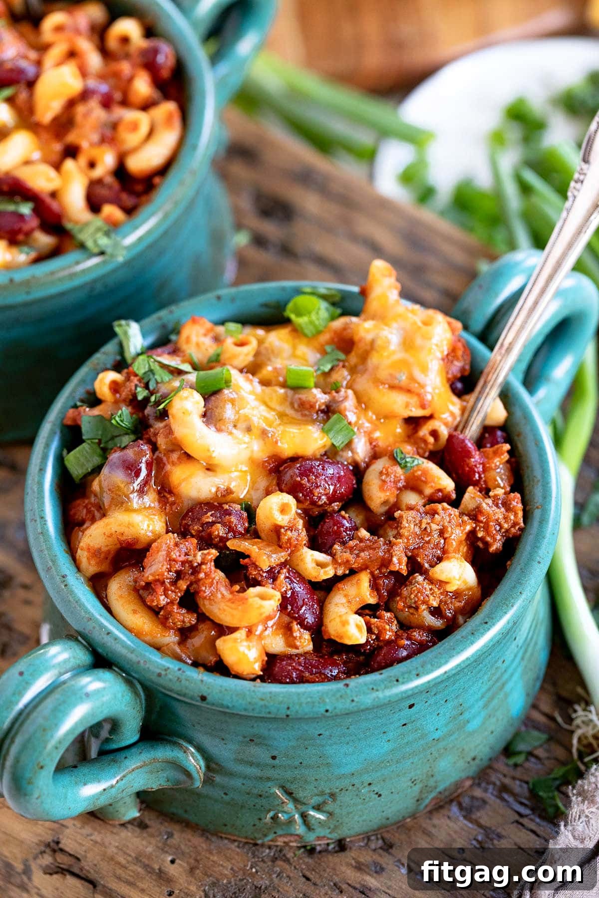 A teal bowl brimming with homemade chili mac, generously topped with shredded cheddar cheese and vibrant sliced green onions, ready to be enjoyed.