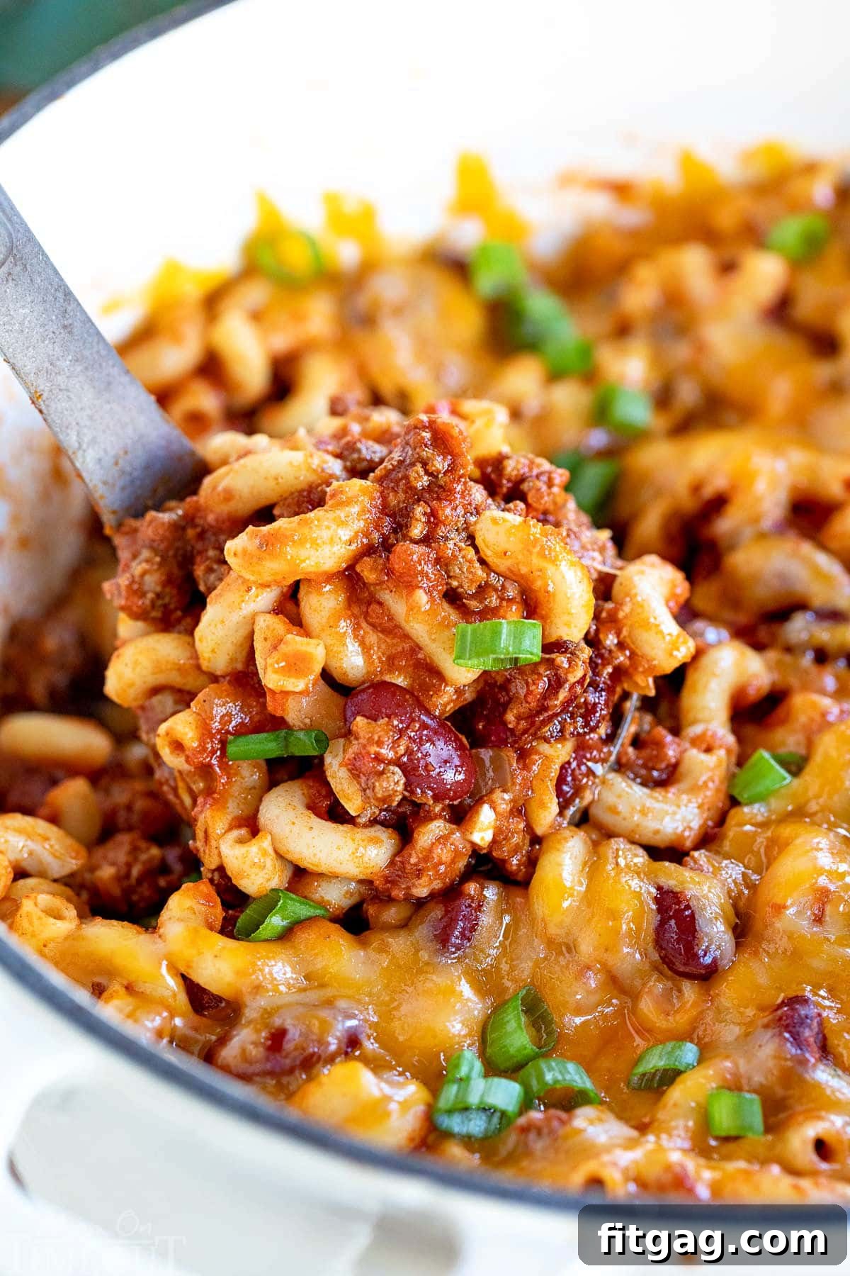 A ladle serving a generous portion of warm chili mac from a pristine white Dutch oven, garnished with fresh green onions.