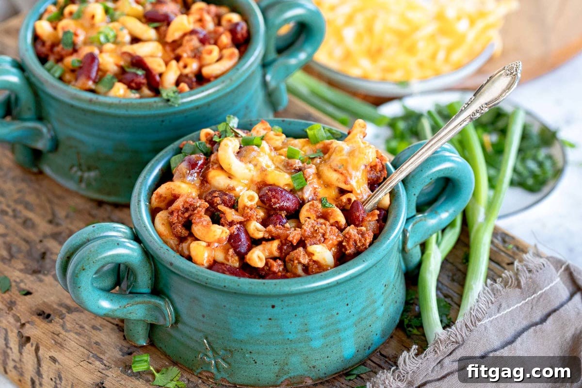 Two inviting bowls of chili mac, generously topped with melted Colby Jack cheese and fresh green onions, resting on a rustic thick wooden board.