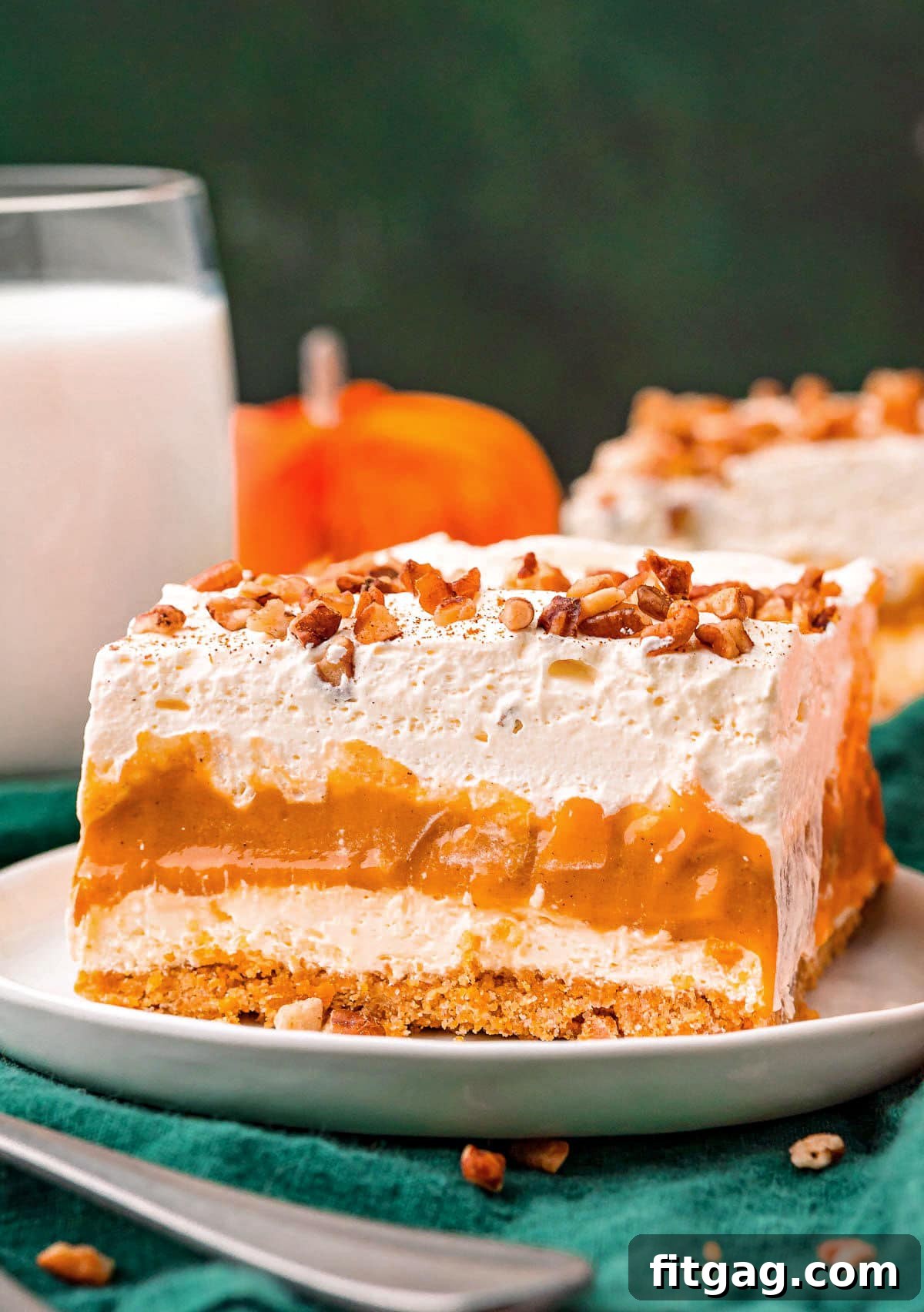 Harvest Pumpkin Bliss 2 A slice of Pumpkin Delight layered dessert on a white plate, garnished with chopped pecans and a dusting of pumpkin pie spice. The distinct four layers are visible, showcasing the graham cracker crust, cheesecake, pumpkin pudding, and whipped topping.